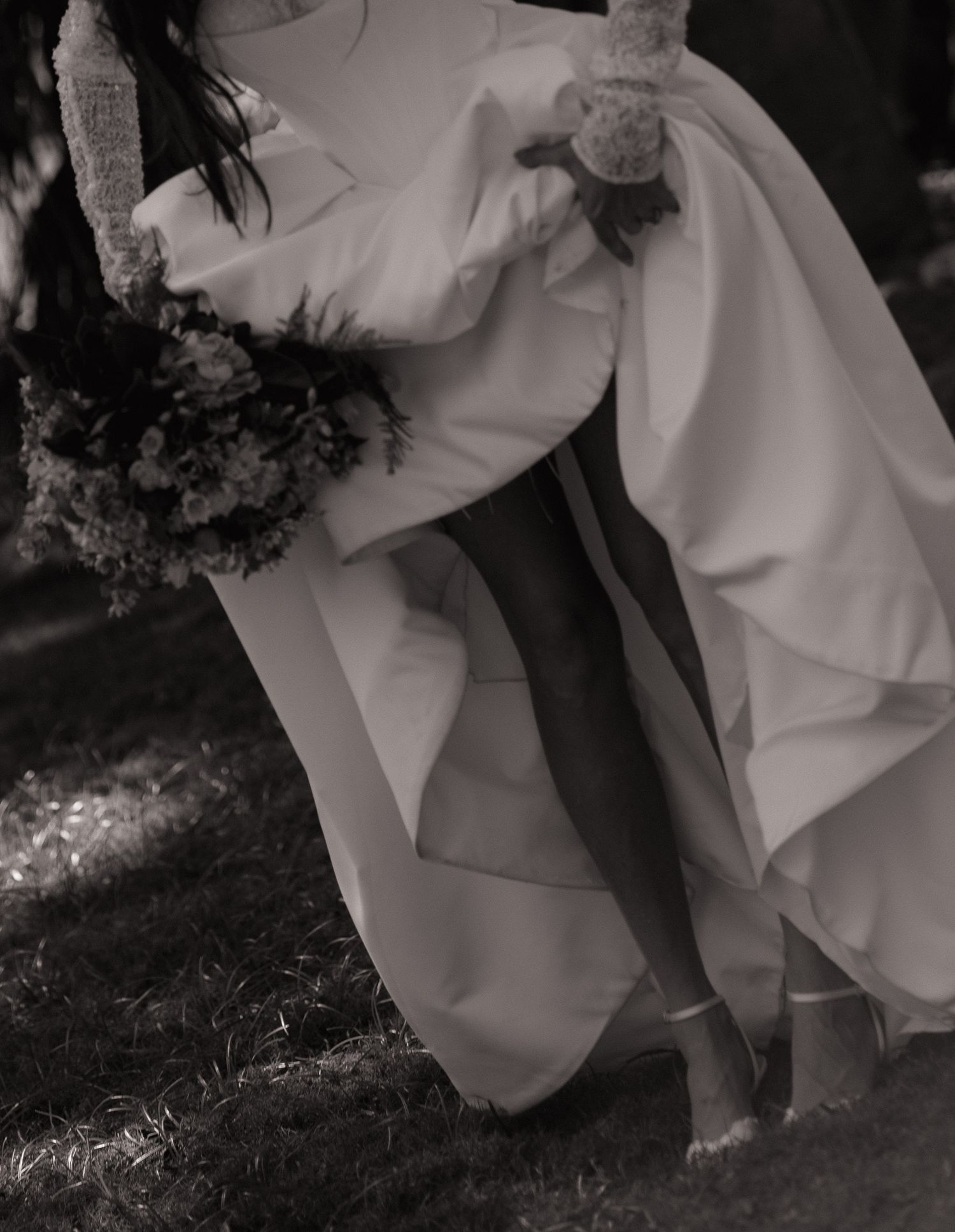 Black and white photo of a bride in a flowing wedding gown holding a bouquet while sitting on a swing outdoors.