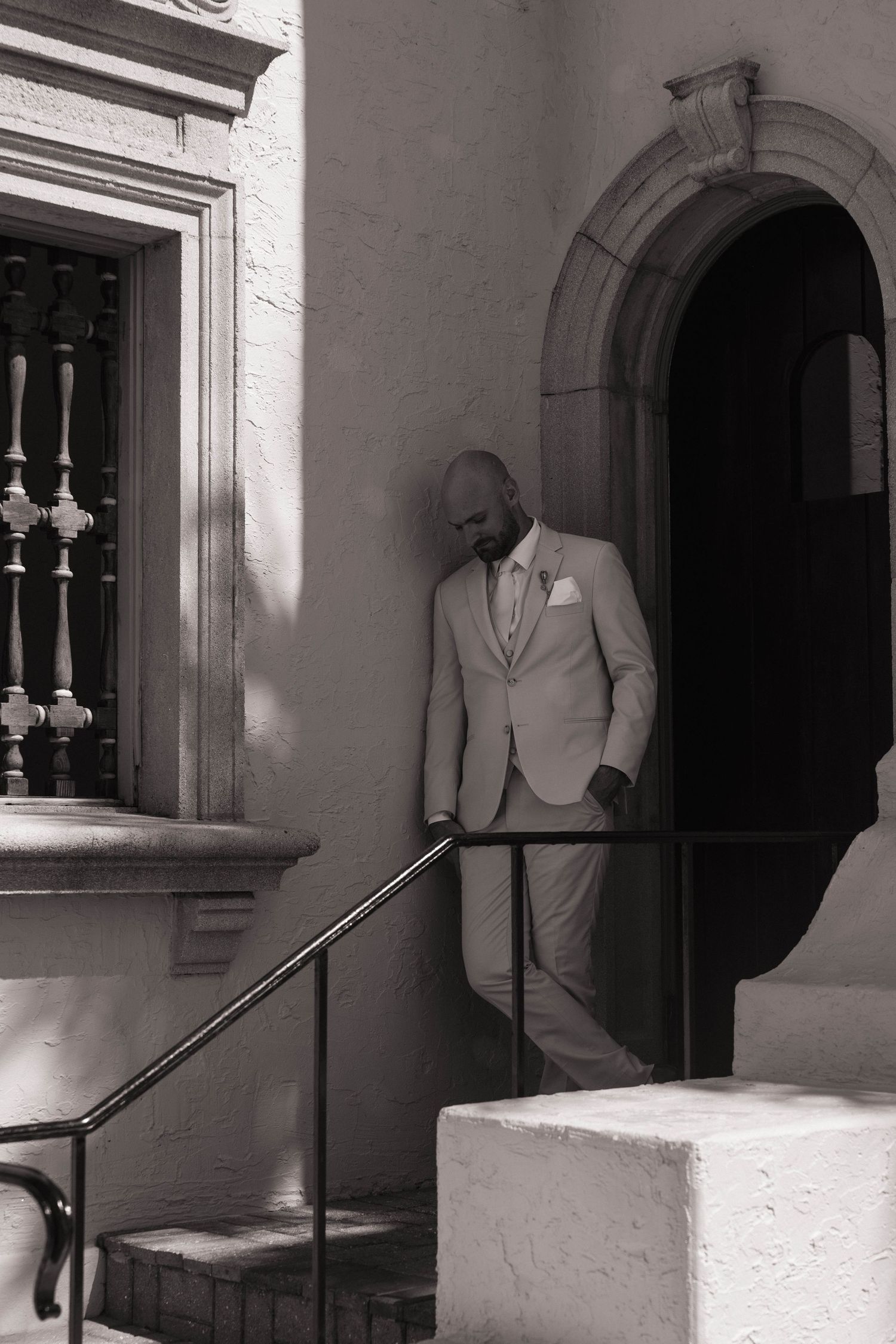 Bald groom in light suit with boutonniere stands at arched church doorway in black and white wedding photo.