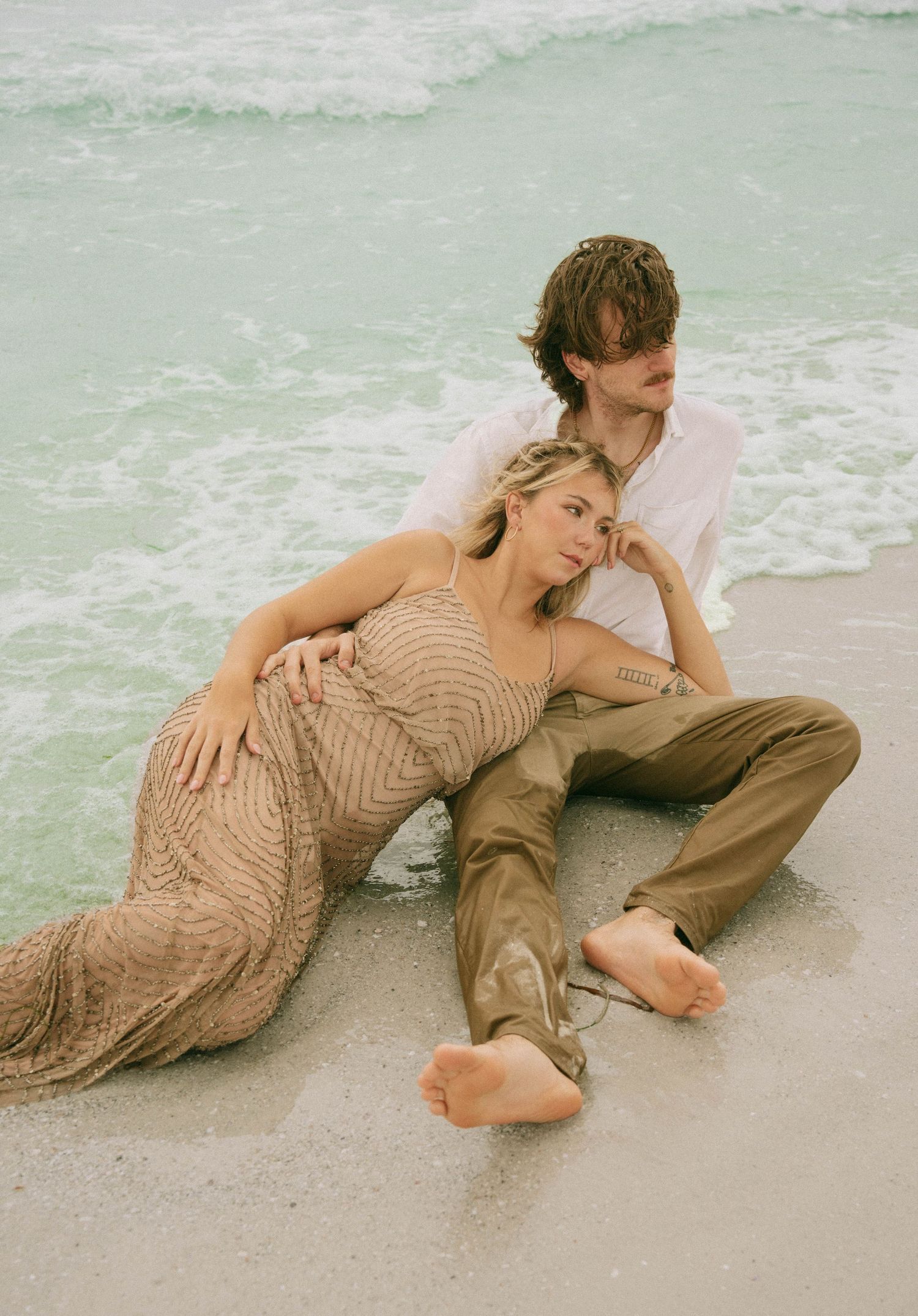 A couple sits together on a sandy beach, the woman in a beaded gown leaning into her partner's embrace.
