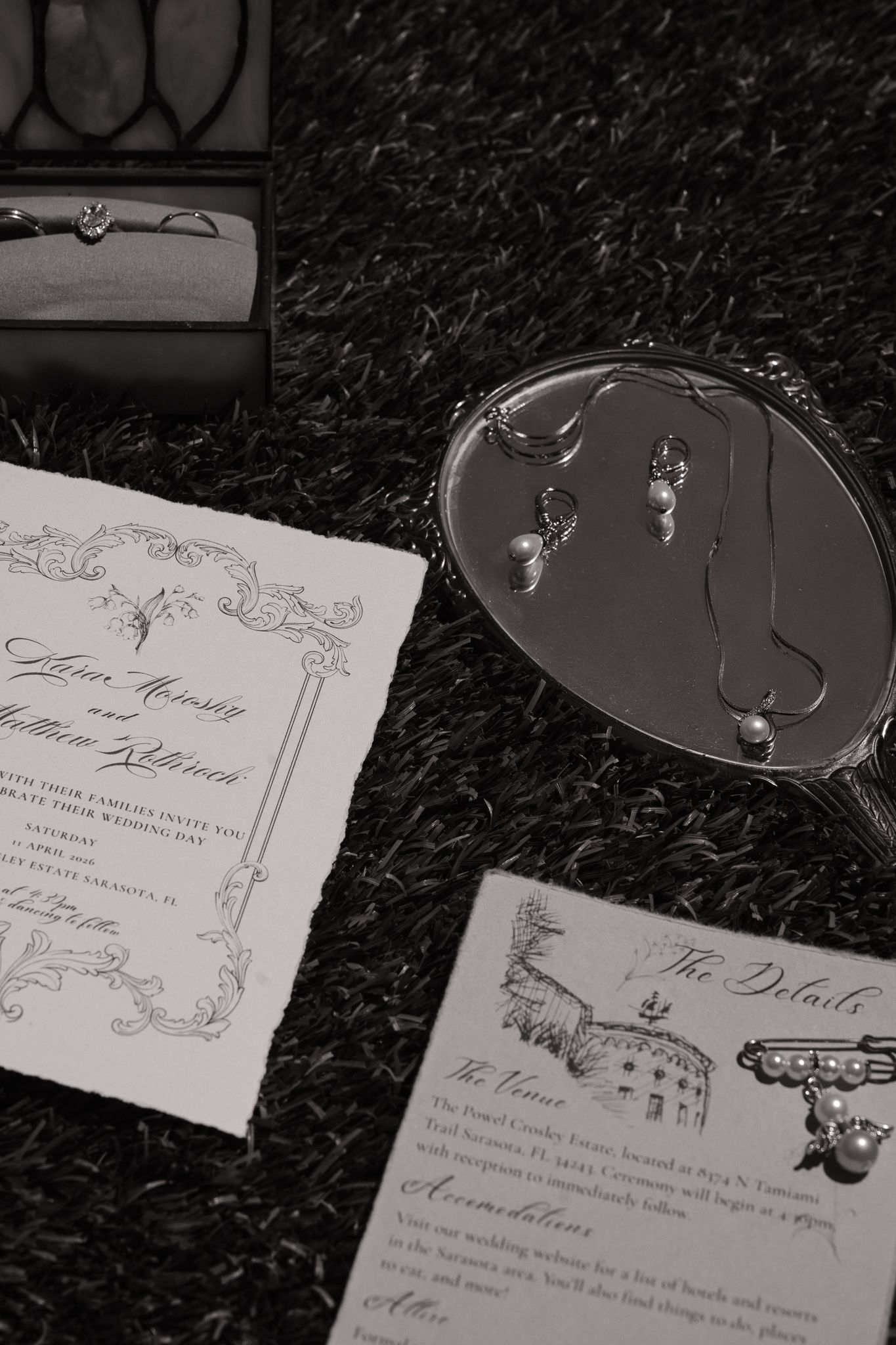 Black and white wedding flat lay with invitation, details card, jewelry, and rings in a decorative box on dark surface.