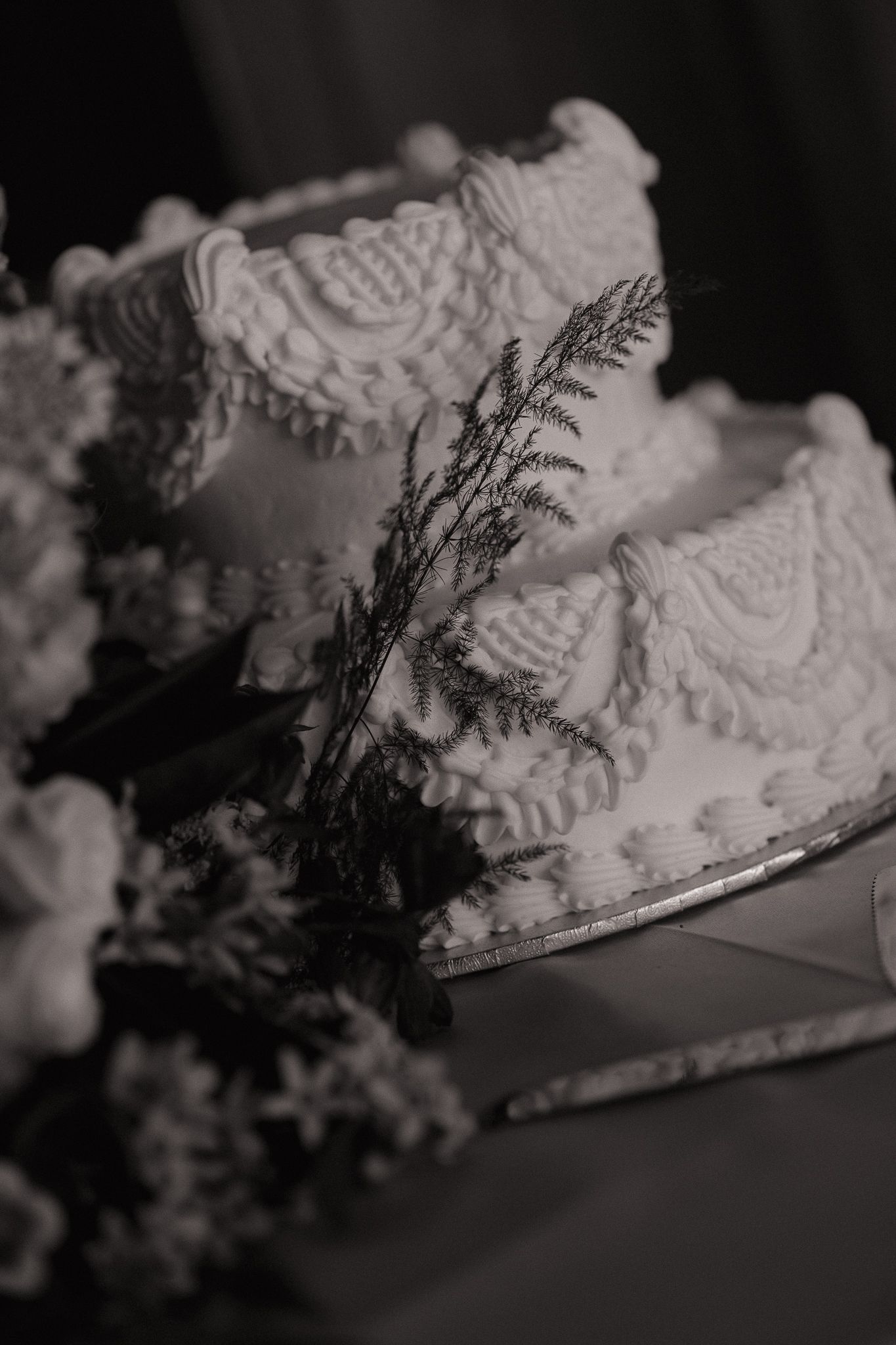 Elegant two-tier white wedding cake with intricate lace frosting details and greenery, captured in black and white.