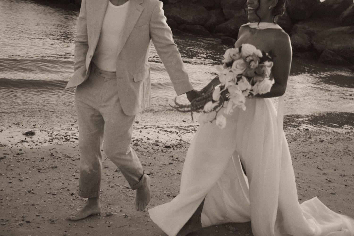 Black and white photo of bride and groom walking hand in hand along a beach.