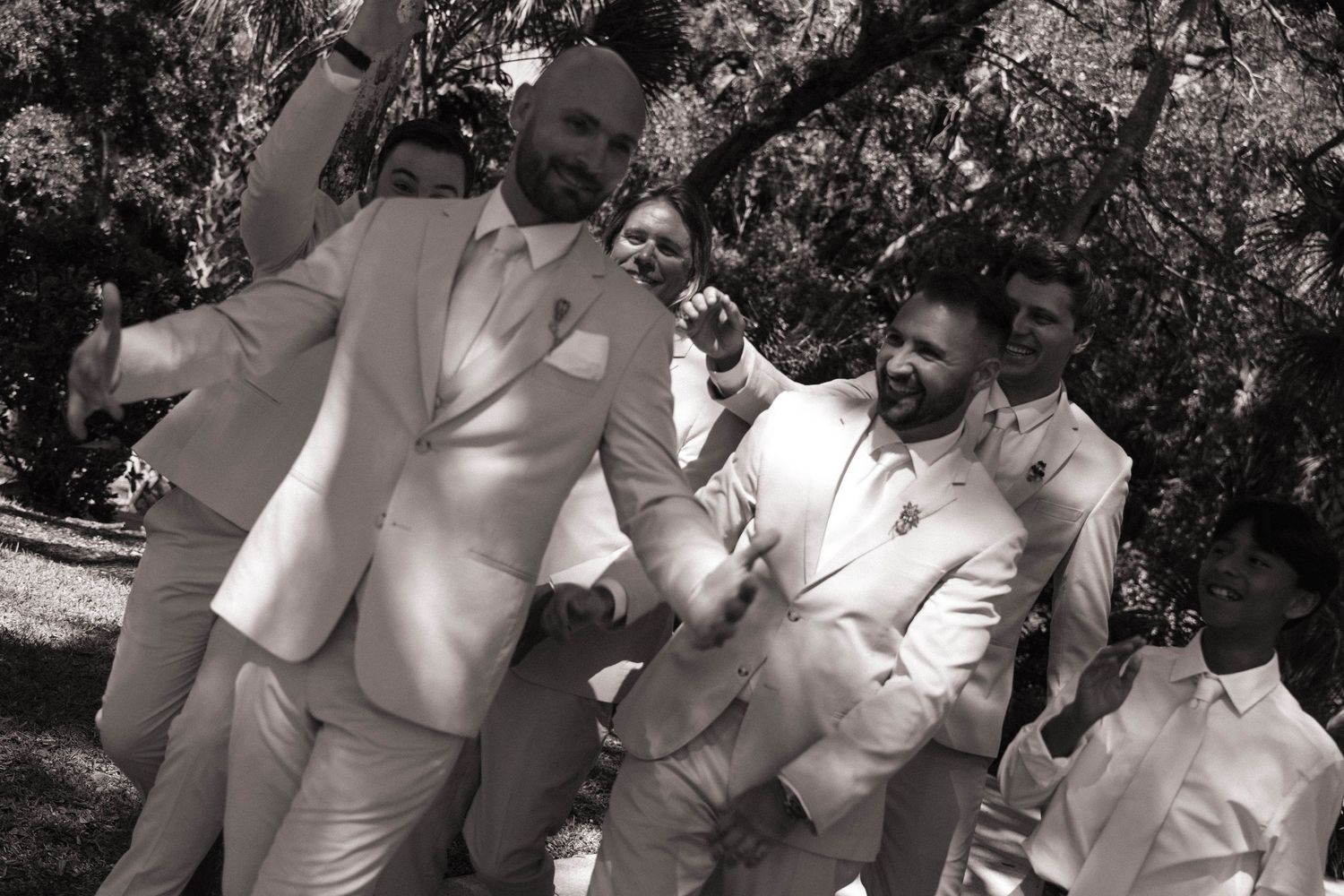 Black and white photo of groom and groomsmen celebrating joyfully outdoors in white suits.