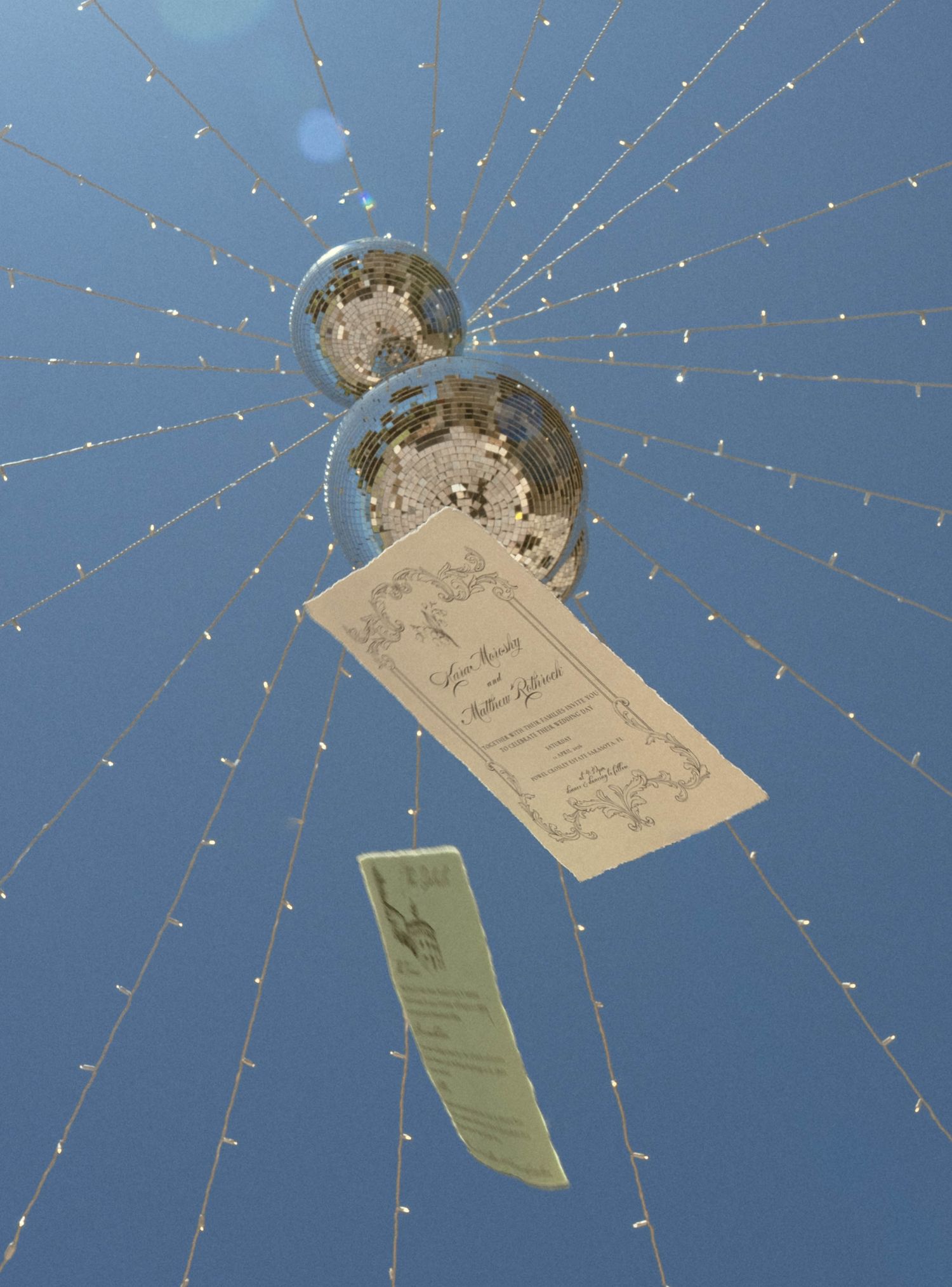Wedding invitation cards hanging from disco balls against a bright blue sky with string lights.