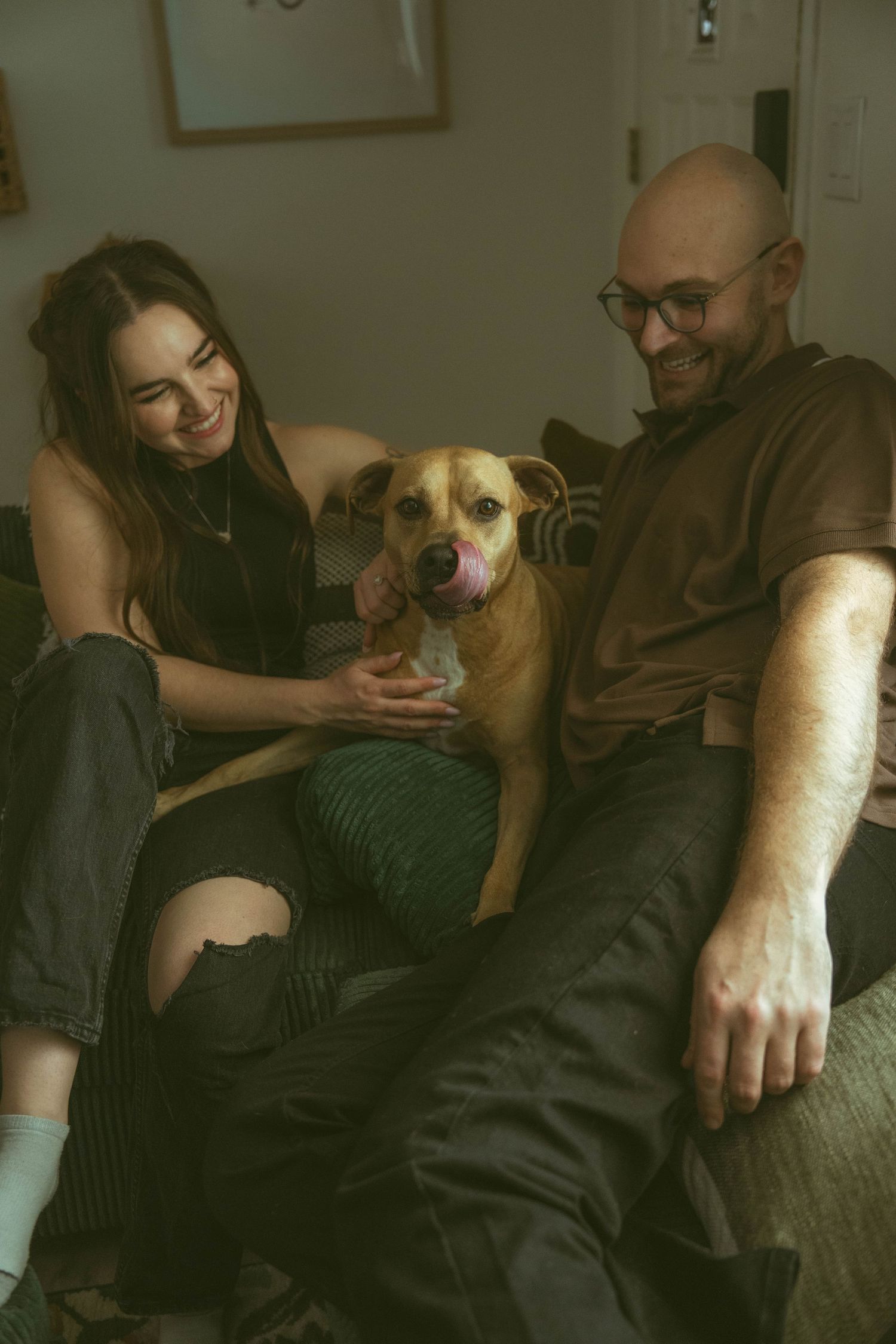 Smiling couple sitting on a couch with their happy tan dog licking its nose.