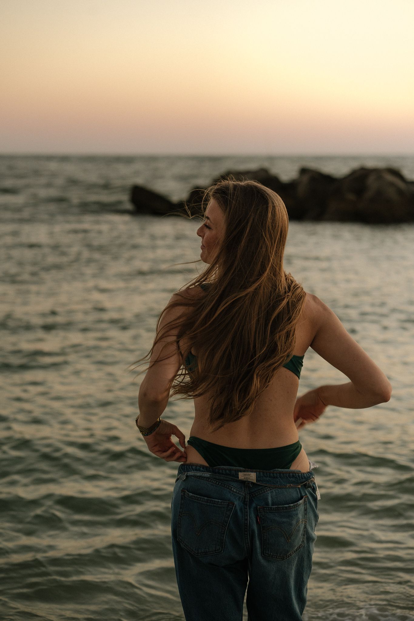 Young woman in green bikini top and jeans stands at rocky ocean beach during golden hour sunset.