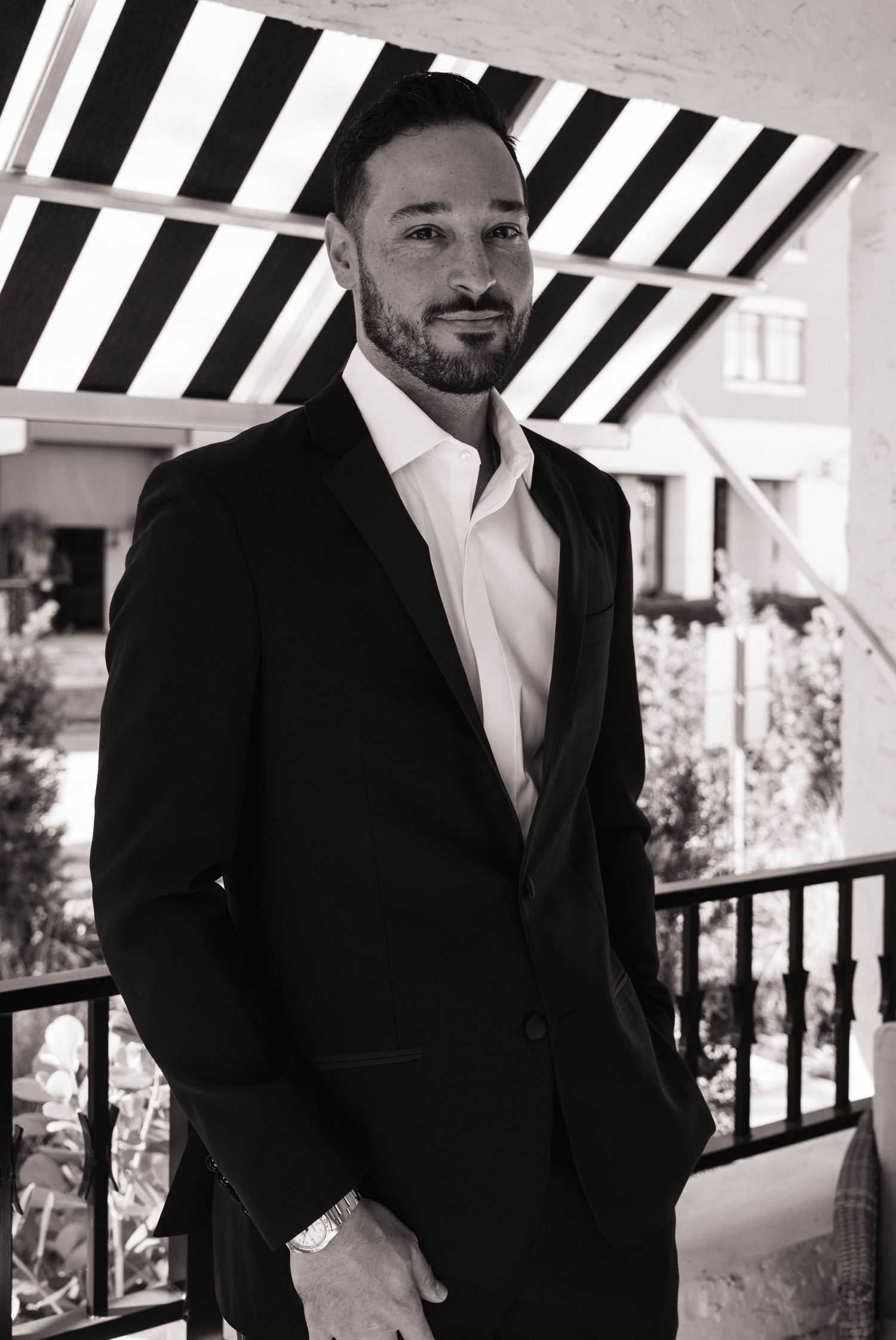 Handsome groom in a black suit poses confidently on a porch in a classic black and white wedding portrait.