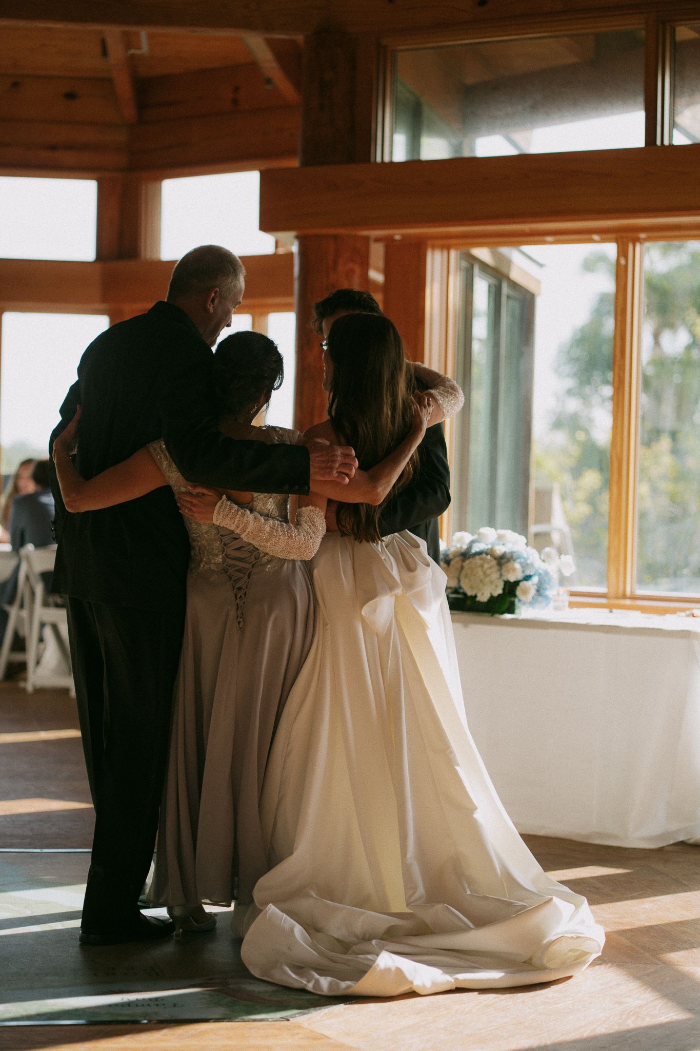 Bride in white gown shares emotional group hug with family members in sunlit wooden wedding venue.