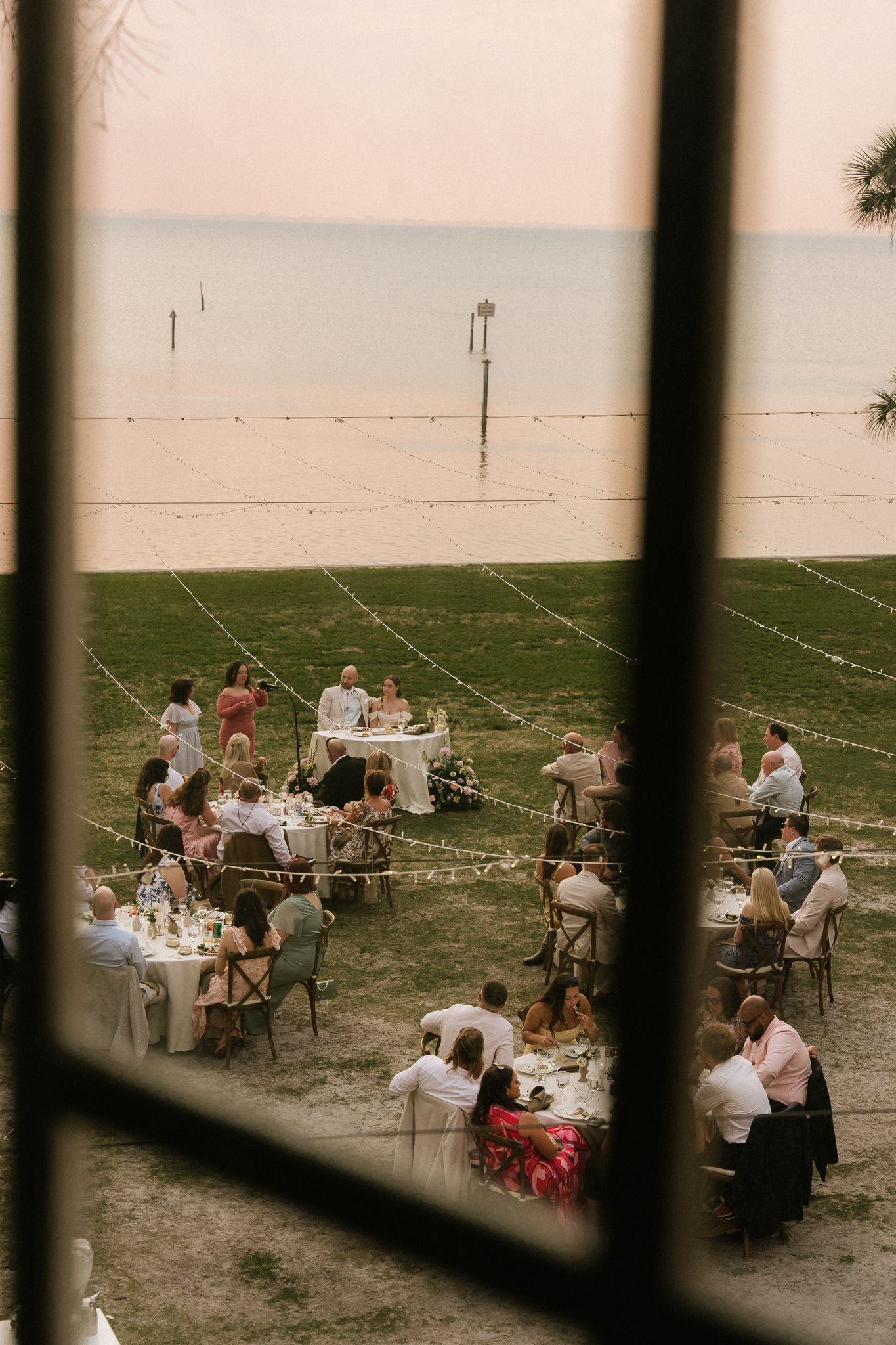 Aerial view of outdoor wedding reception dinner by the ocean at sunset with string lights.