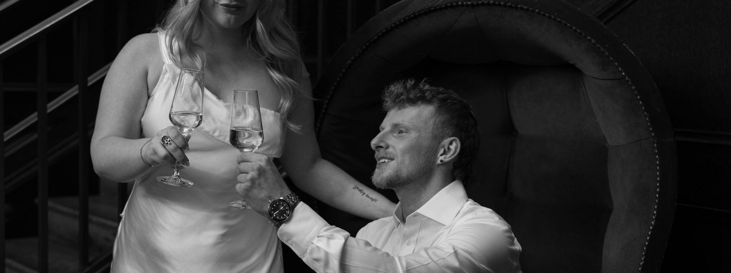 Black and white photo of a couple sharing champagne, woman in white dress standing while man sits smiling up at her.