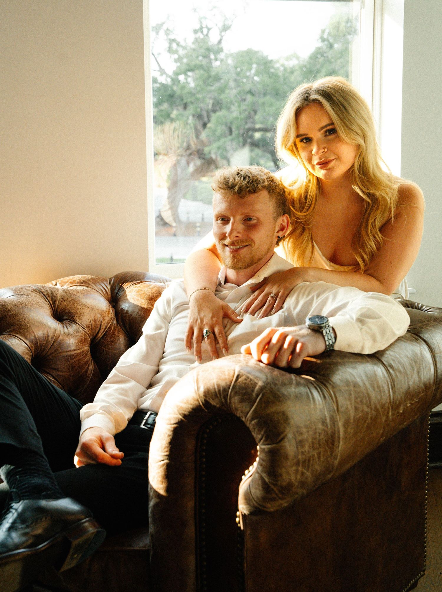 Couple posing on leather sofa near window, woman in yellow dress embracing man in white shirt.