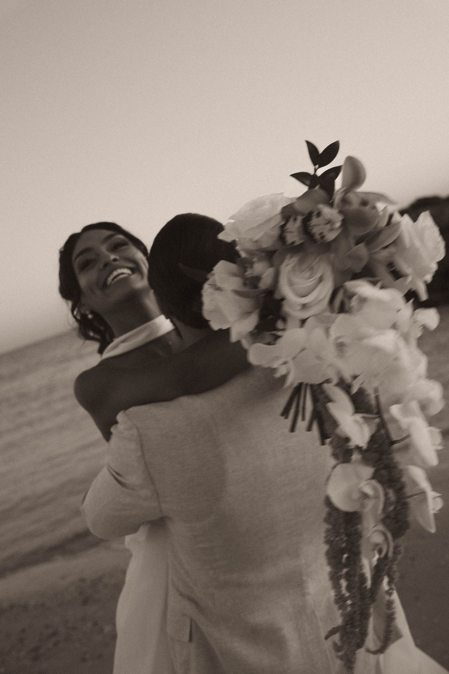Black and white photo of a joyful bride hugging groom while holding a lush floral bouquet on a beach.