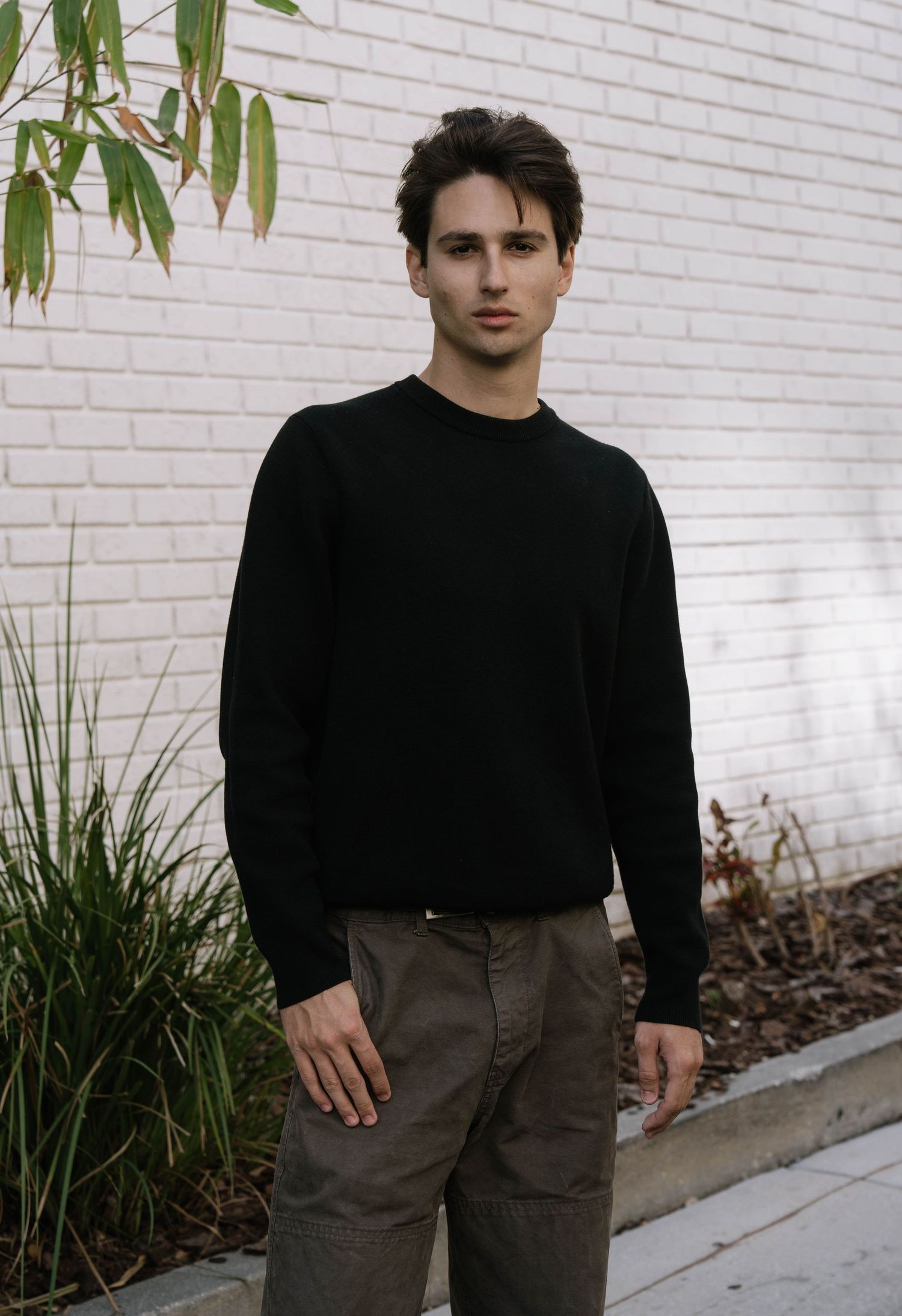 Young man in a black crewneck sweater standing against a white brick wall outdoors.