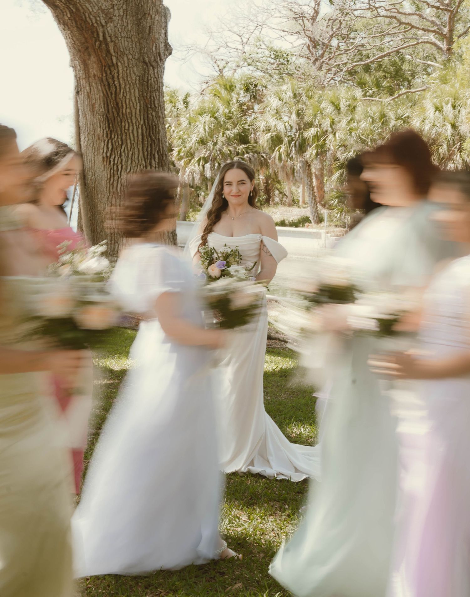 Bride in white off-shoulder gown surrounded by blurred bridesmaids holding bouquets in outdoor garden.