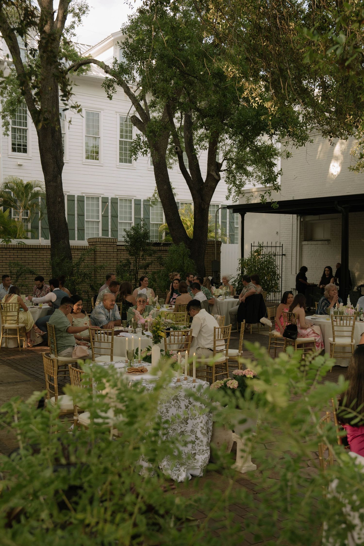 Elegant outdoor wedding reception with guests seated at gold chiavari chairs under lush trees beside a white historic venue.