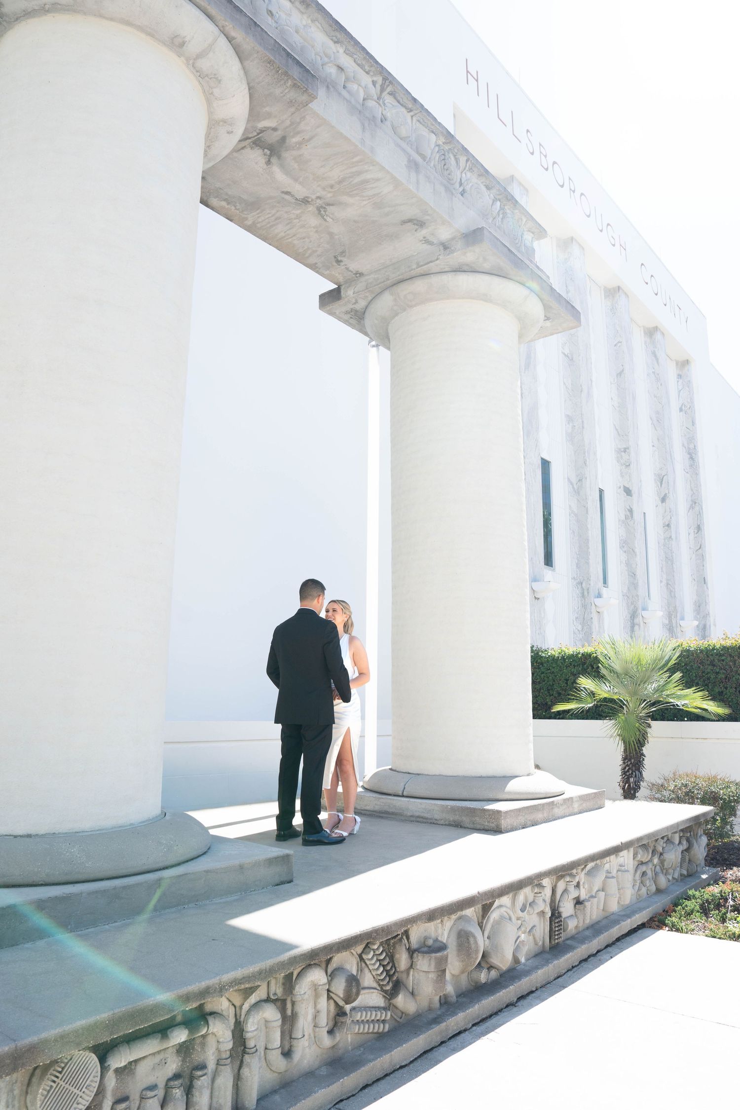 Wedding couple standing between tall white columns outside classical courthouse building in sunlight.