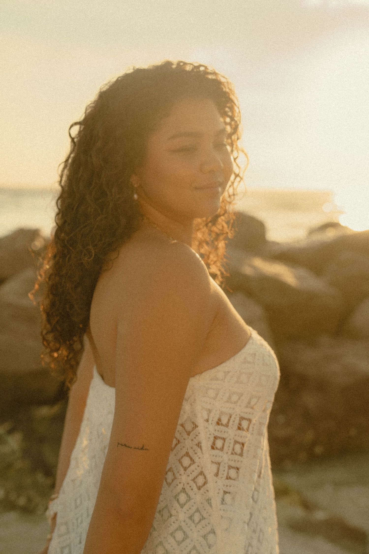 Woman with curly hair in white lace dress smiling at beach during golden sunset.