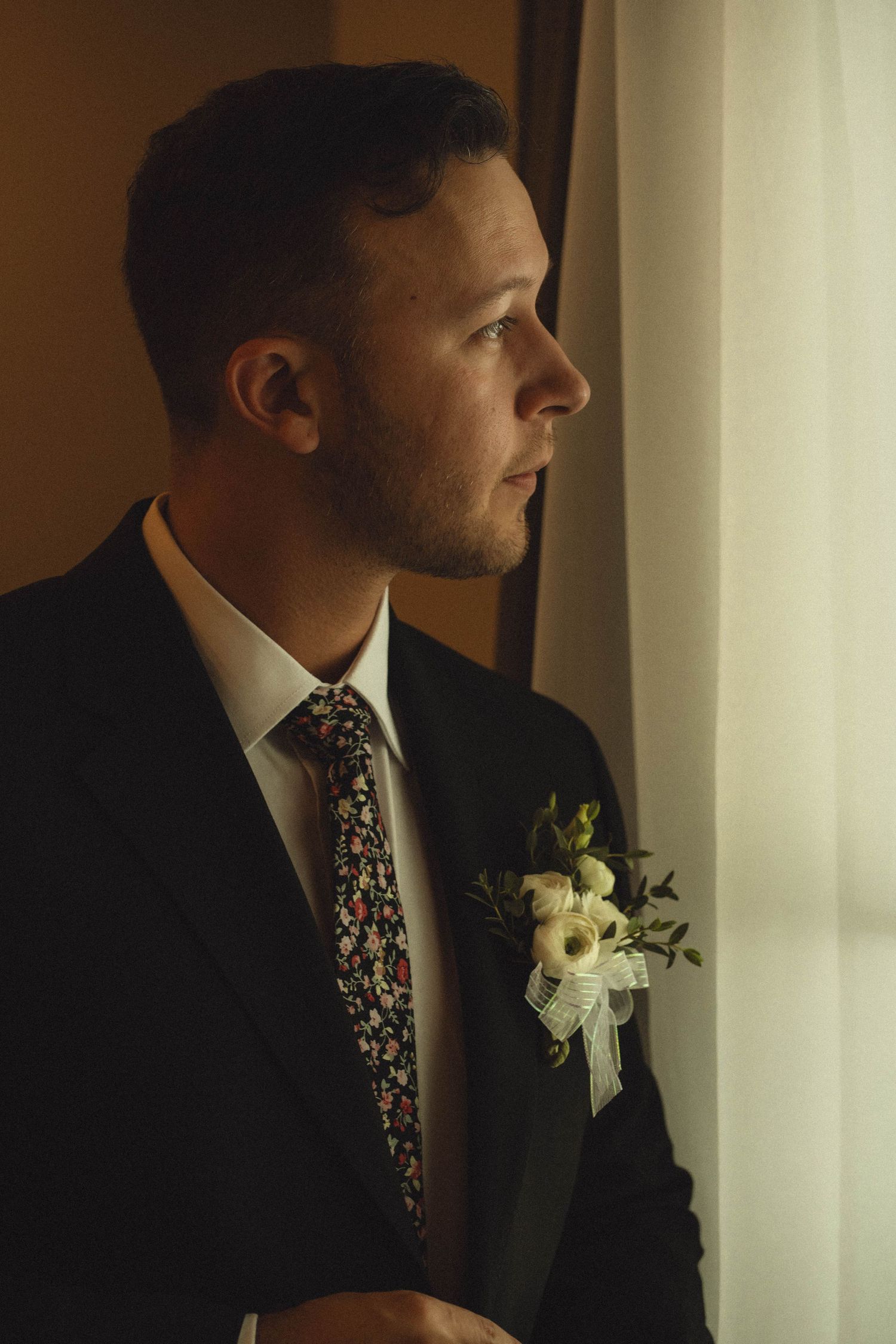 Groom in dark suit with floral tie and boutonniere gazes thoughtfully out window before wedding.