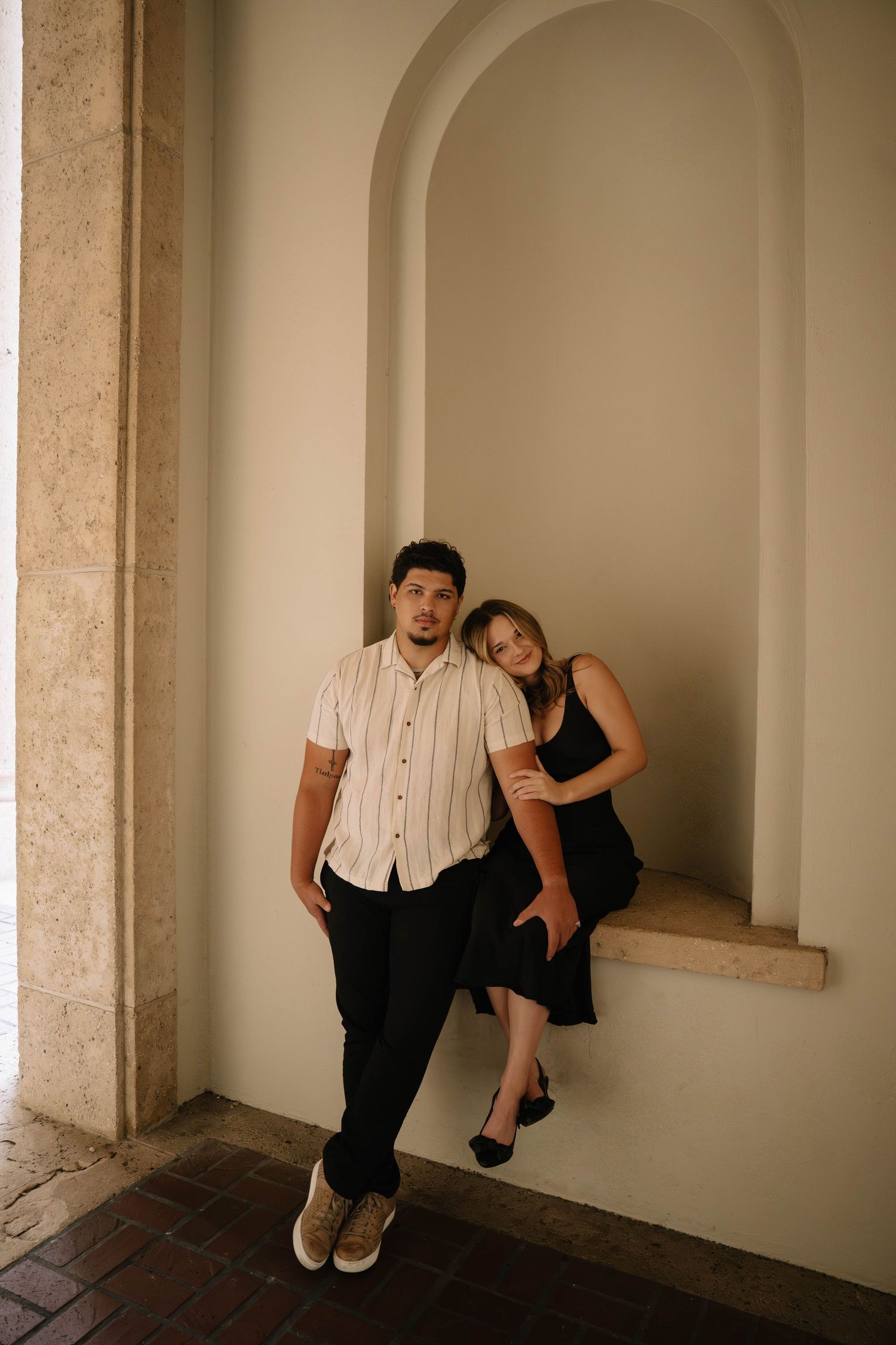 Couple posing in an elegant arched alcove, woman in black dress leaning on mans shoulder.