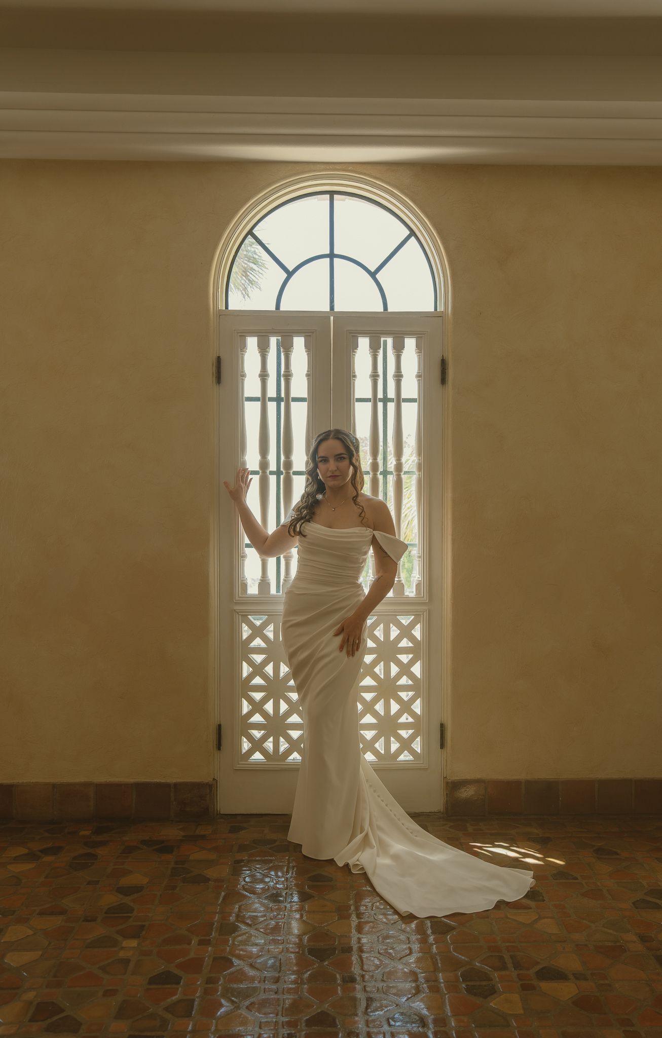 Bride in elegant white satin off-shoulder gown standing before arched French doors in a sunlit room.
