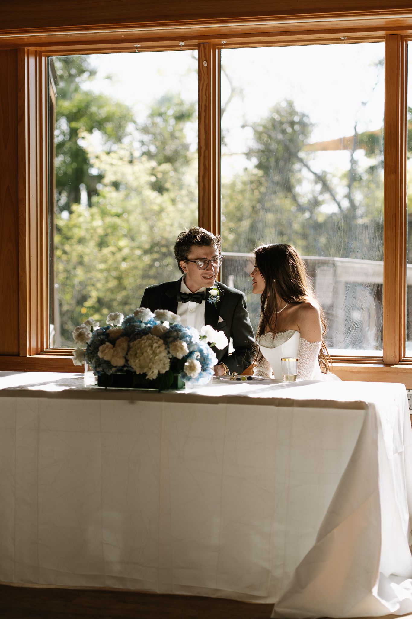 Bride and groom sit together at sweetheart table adorned with blue hydrangea centerpiece by sunny window.