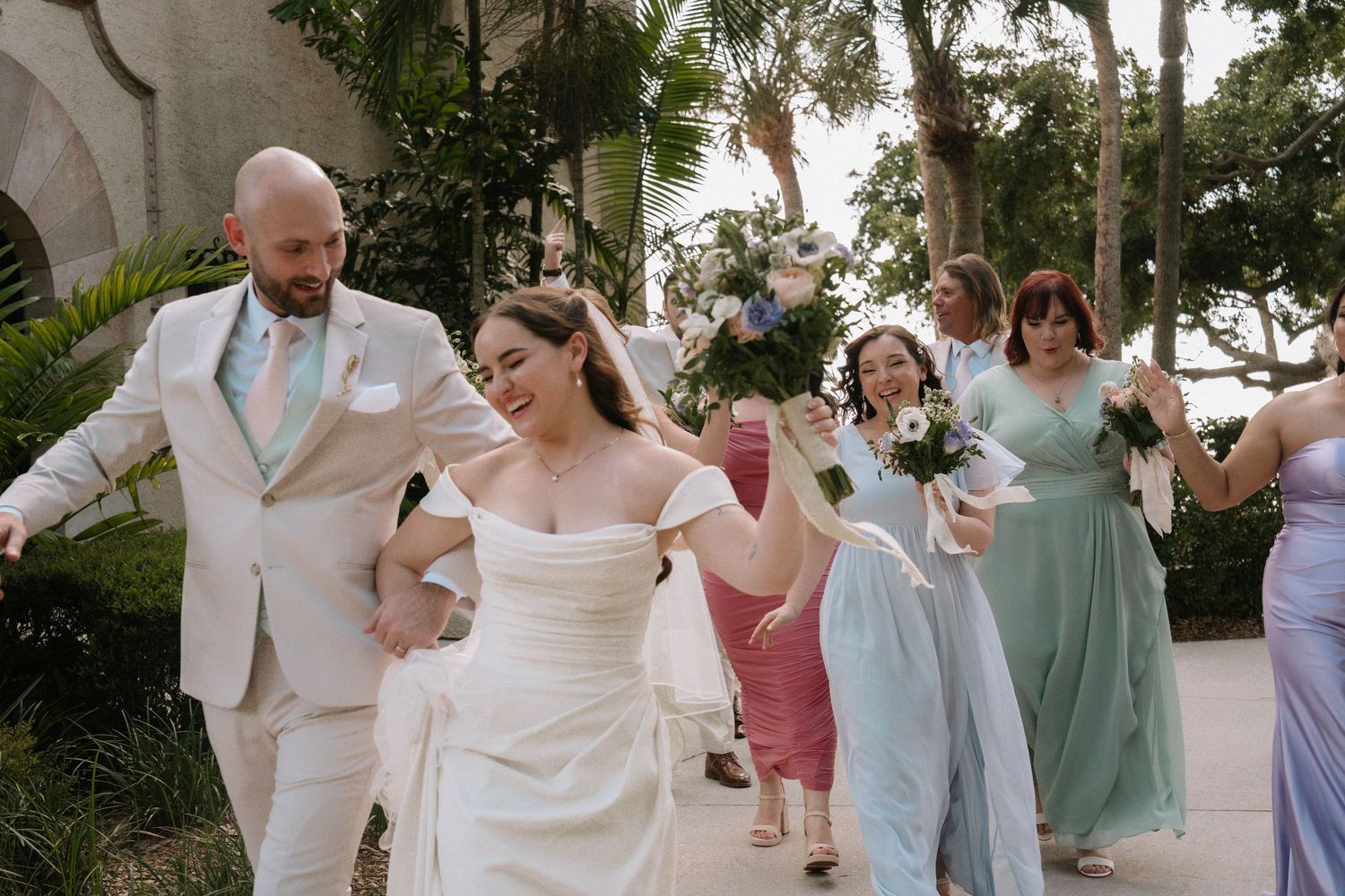Joyful wedding party walks outdoors, bride in white gown with groom in light suit, bridesmaids in sage green dresses.