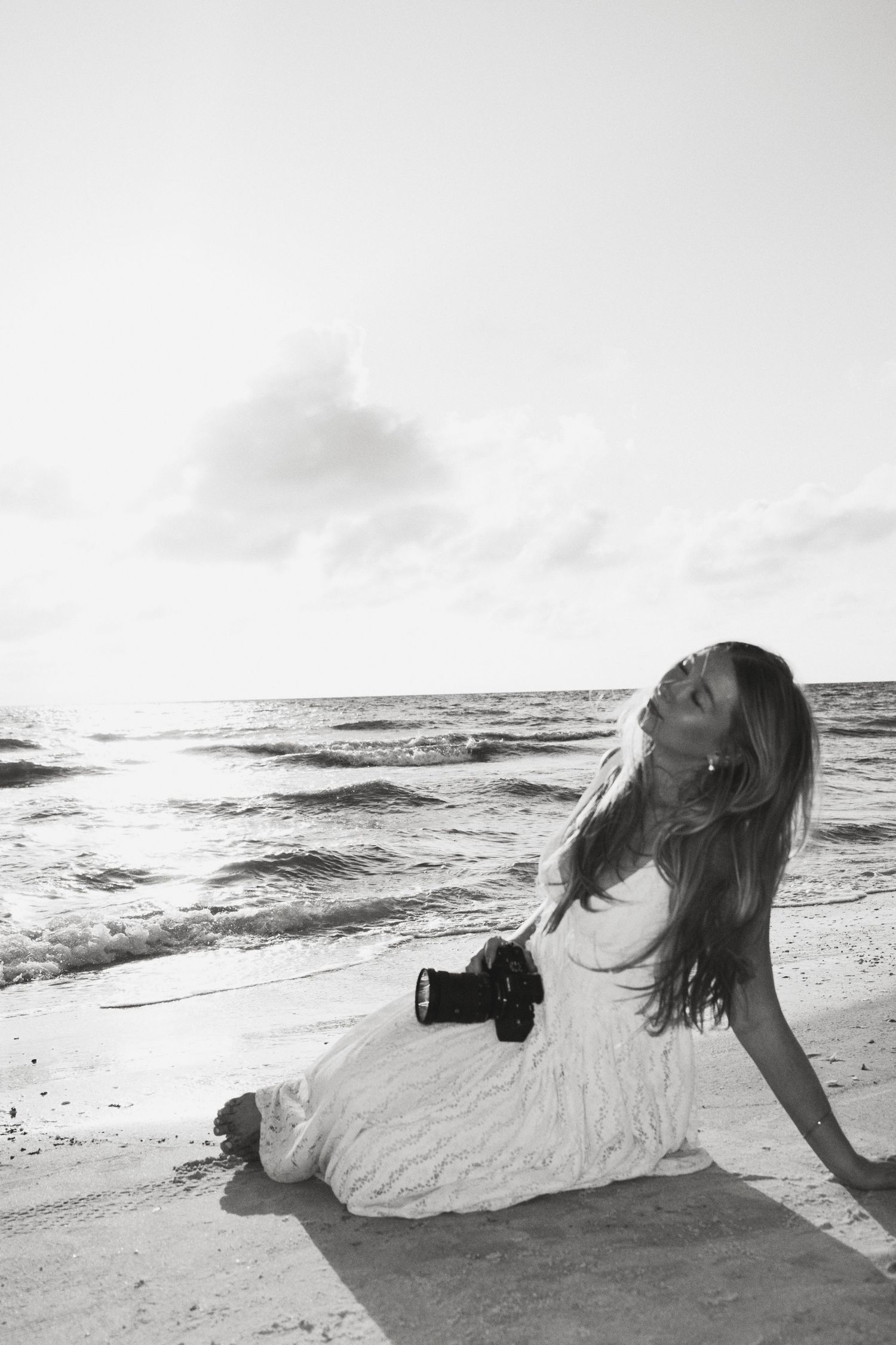 Black and white photo of a woman in a white dress sitting on a beach holding a camera, with ocean waves behind her.