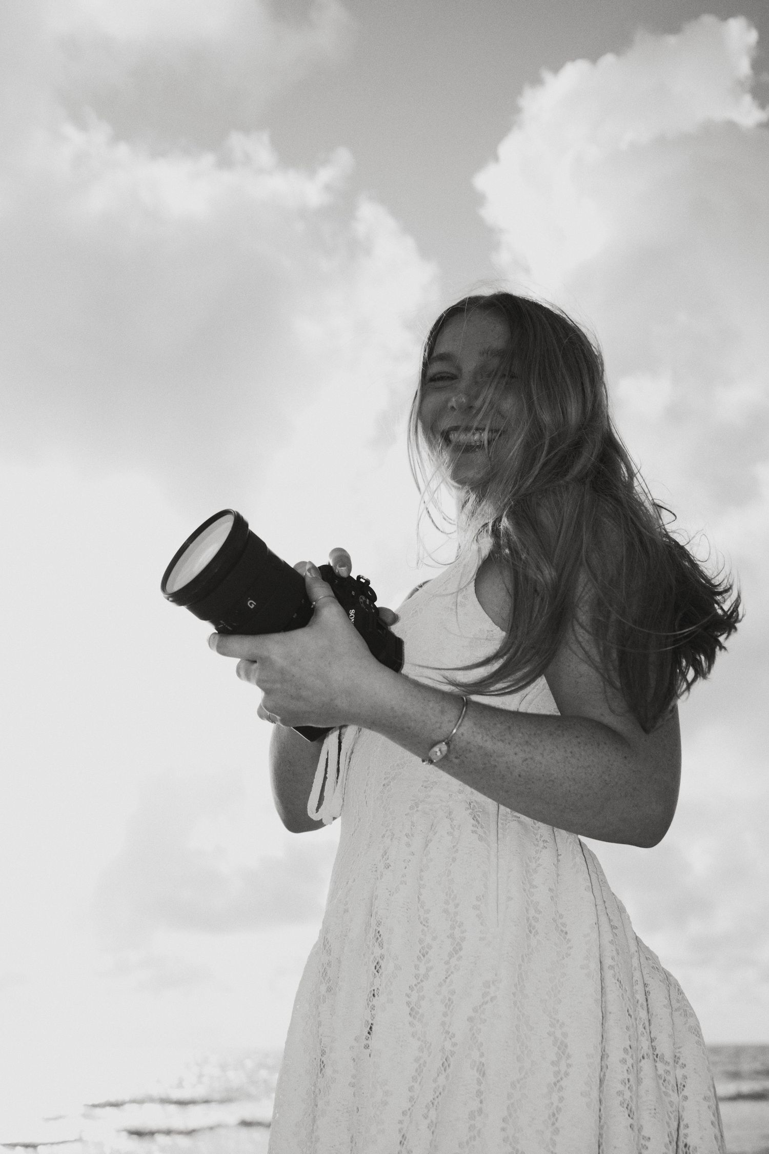 Black and white photo of a smiling young woman in a white dress holding a camera outdoors with cloudy sky background.