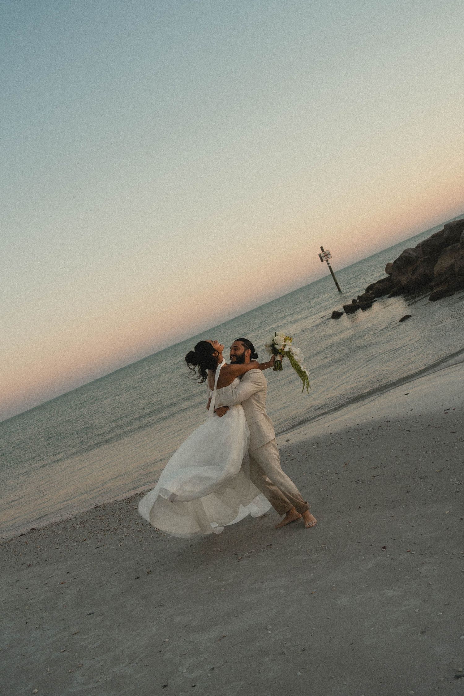 Bride and groom dance joyfully on beach at sunset, bride in flowing white gown holding bouquet.