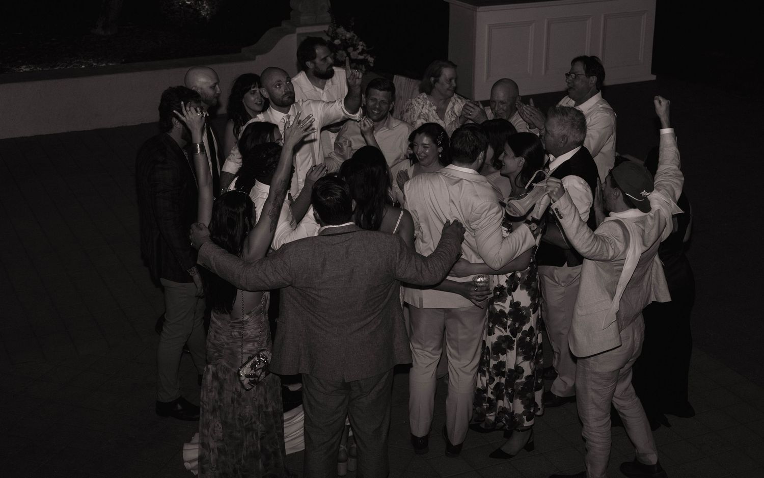 Black and white photo of wedding guests gathered in a joyful group celebration at night.