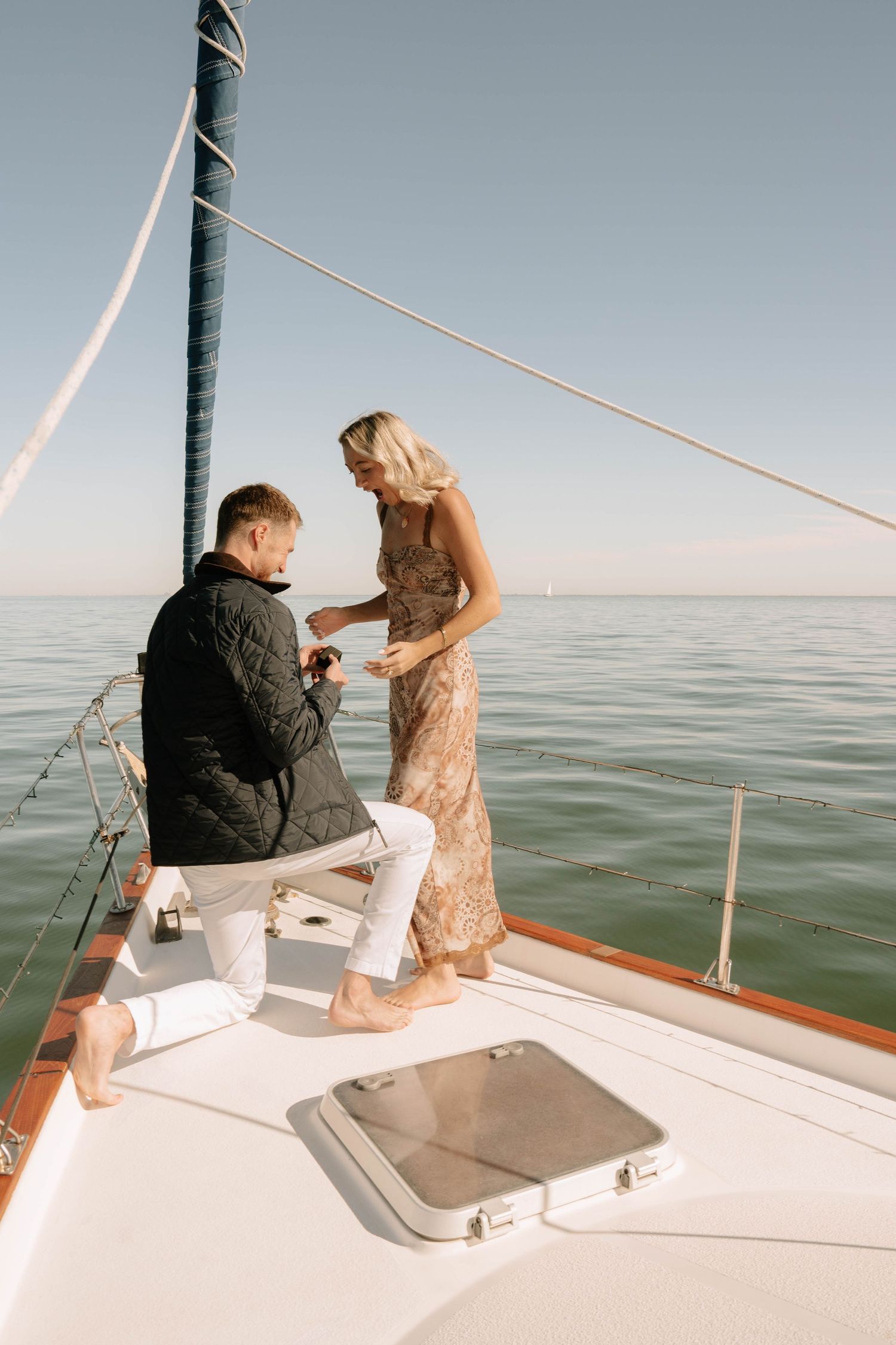 Man proposing on one knee to woman on a sailboat deck with calm ocean backdrop.