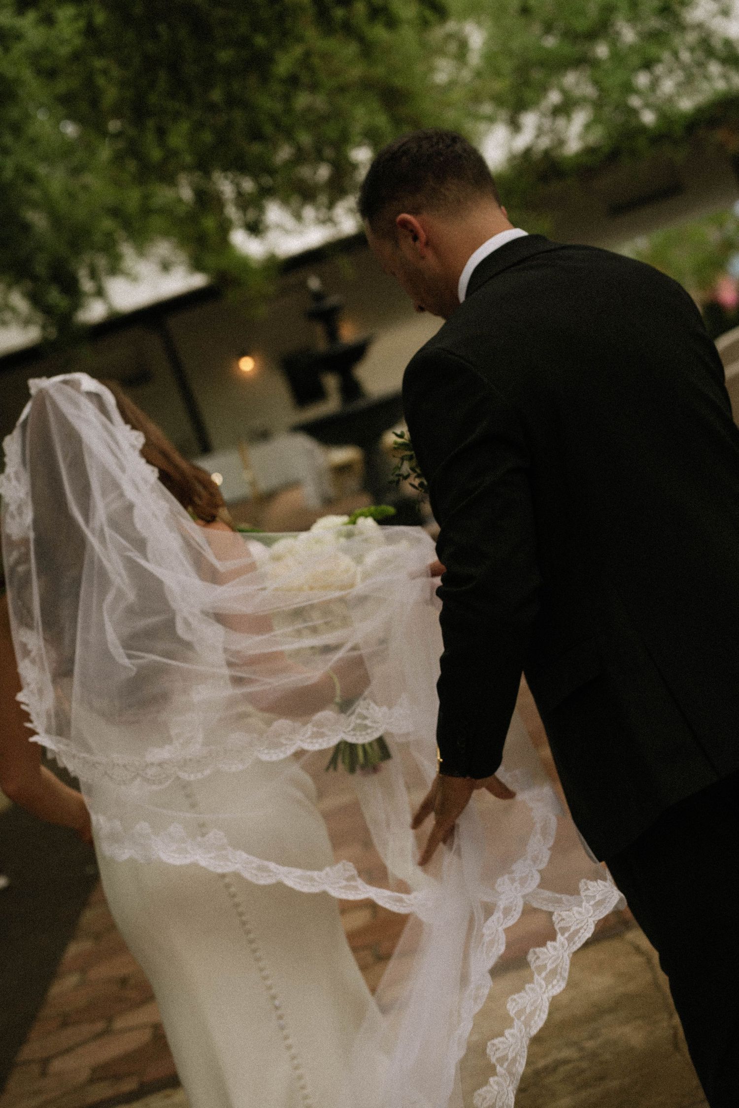 Groom in black suit adjusts bride's lace-trimmed cathedral veil outdoors near a fountain.