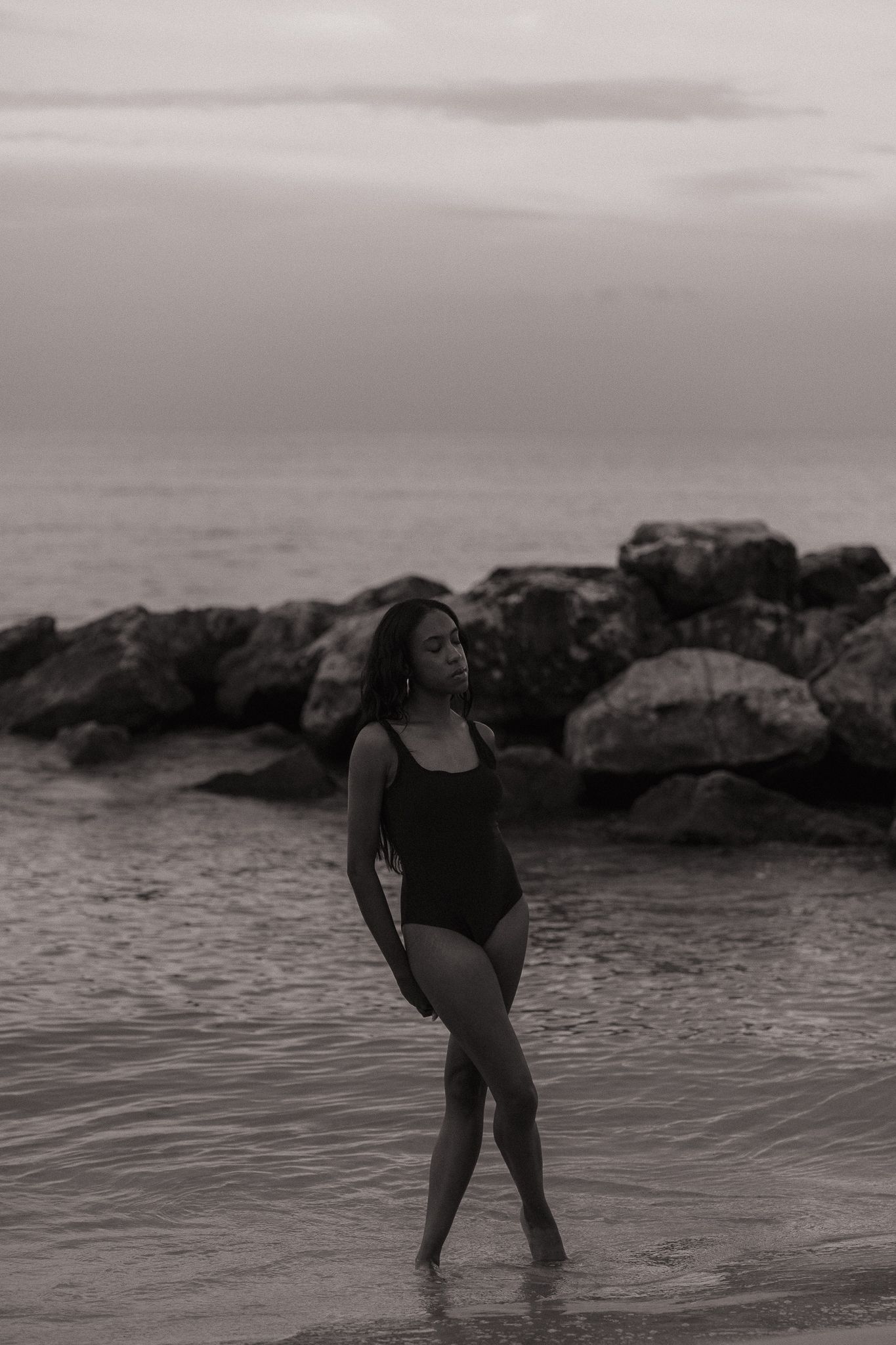 Black and white photo of a woman in a swimsuit standing in shallow ocean water near rocky shore on an overcast day.