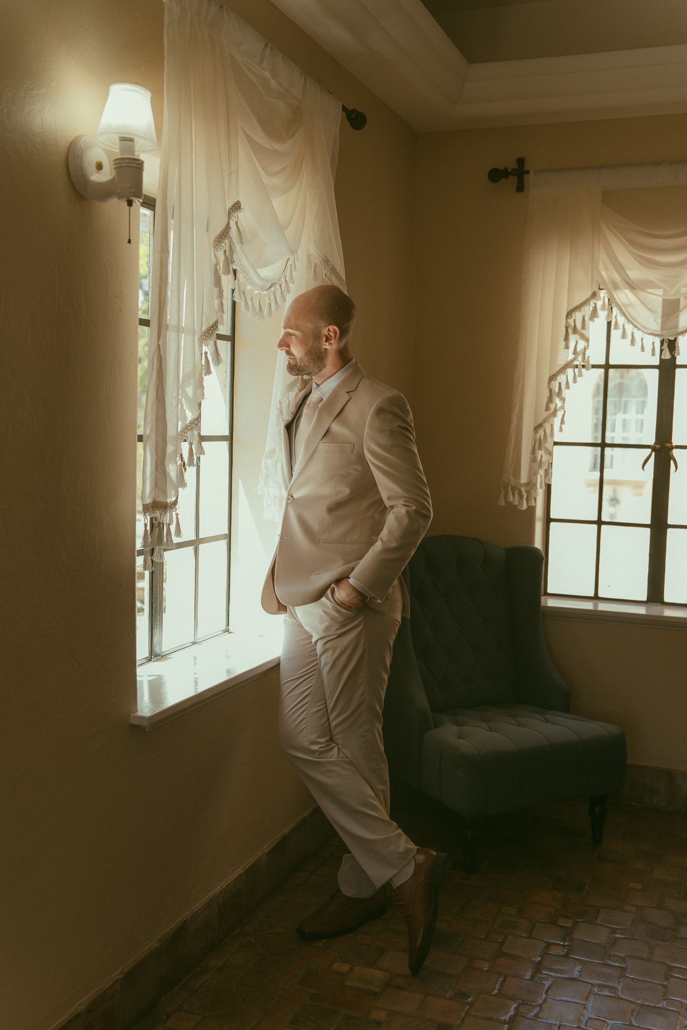 A well-dressed bald man in a beige suit stands by a window in an elegant room, gazing outside thoughtfully.