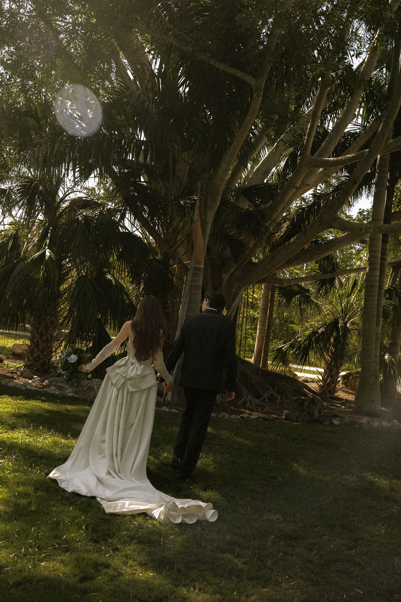 Bride and groom walking hand in hand through lush garden with large trees on their wedding day.