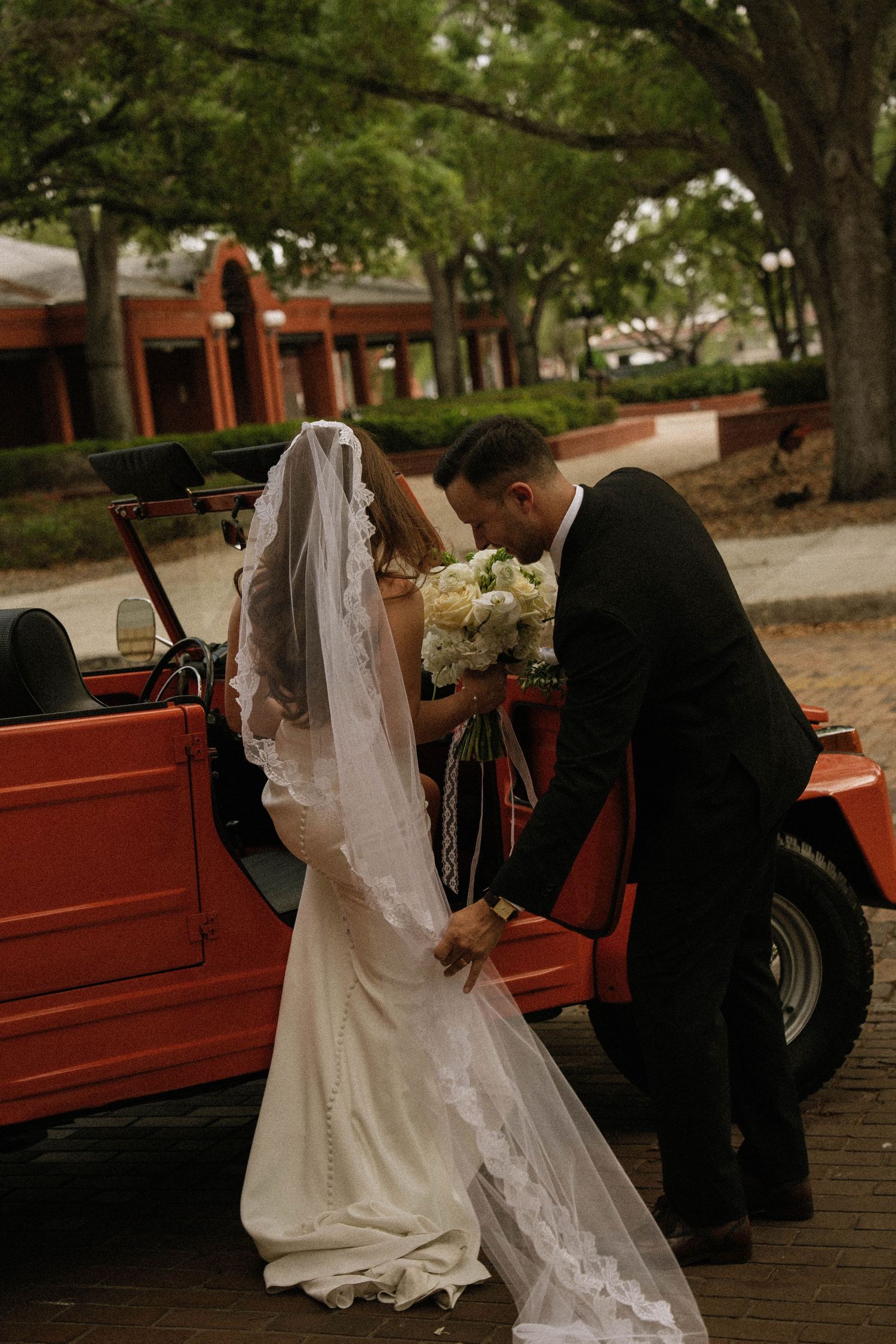 A groom in a black suit helps his veiled bride exit a red vintage vehicle while holding her bouquet.