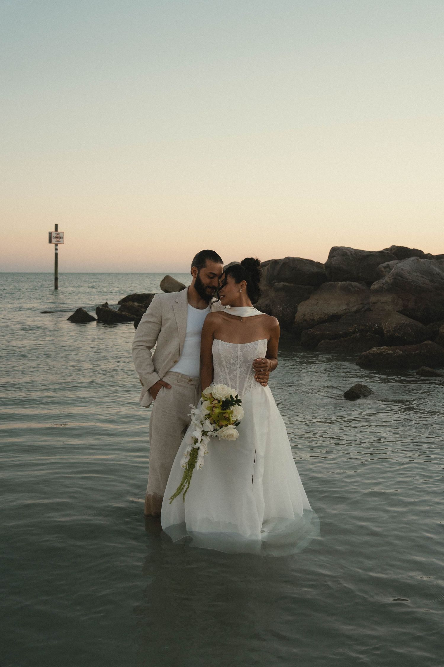 Bride and groom stand romantically in ocean water at sunset, surrounded by rocks and calm sea.