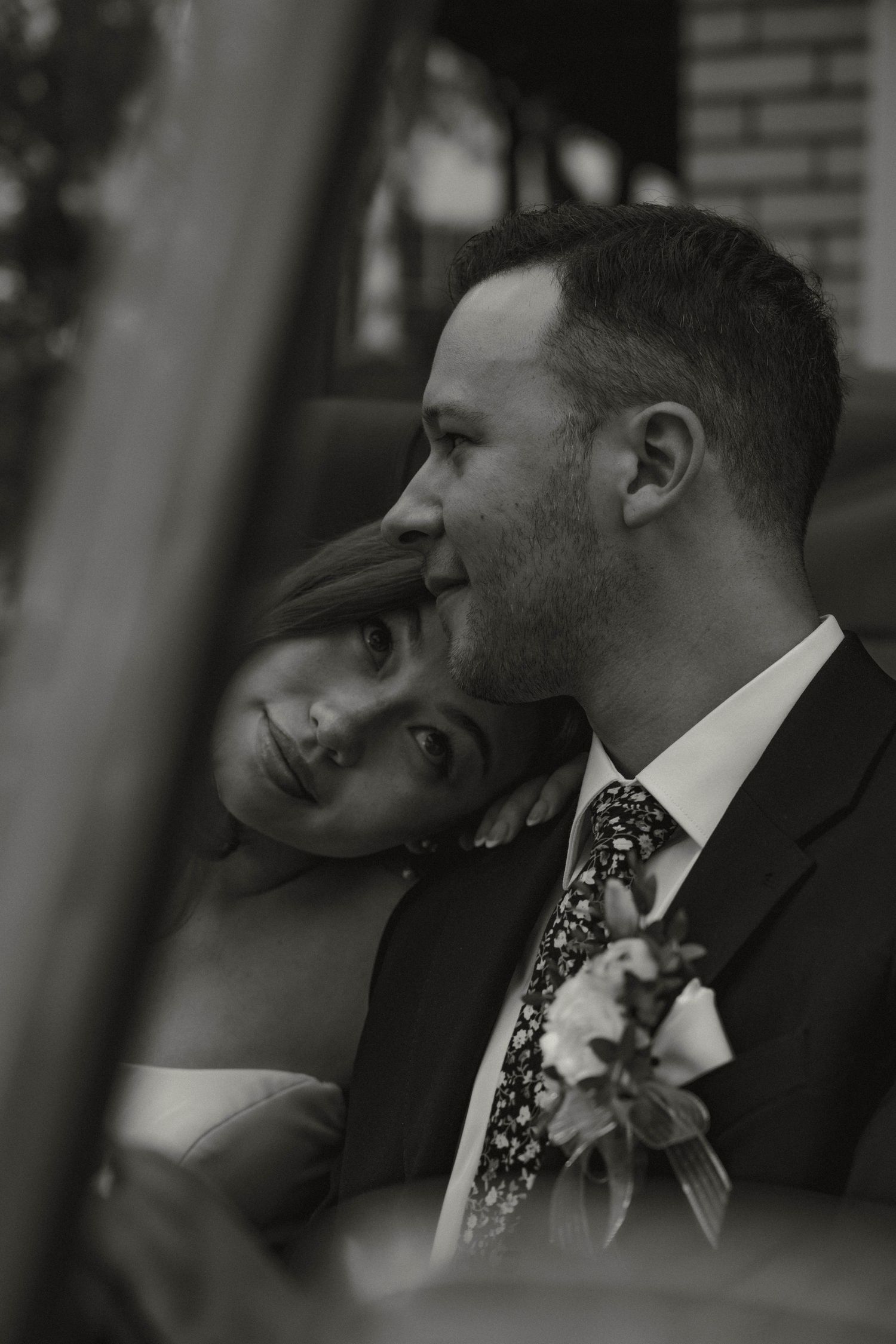 Black and white romantic portrait of bride resting head on groom's shoulder inside a vintage car.