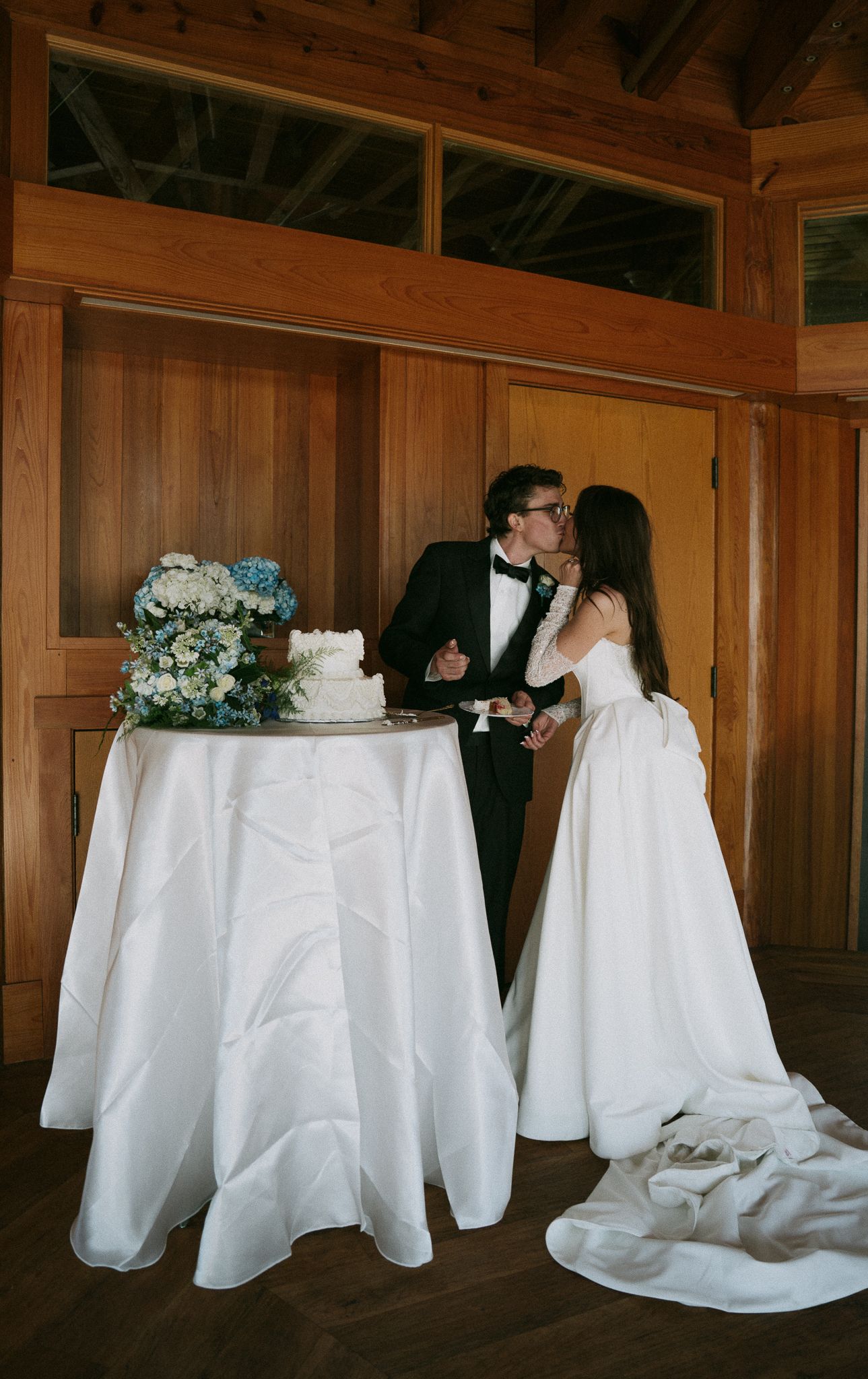 Bride and groom kiss while cutting their white wedding cake adorned with blue hydrangeas.