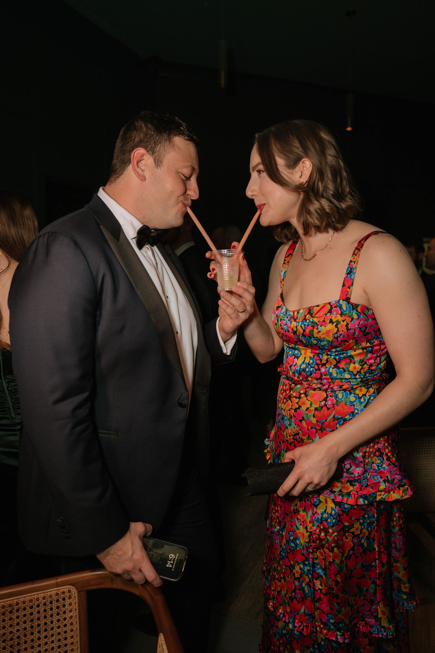 Couple in formal wedding attire playfully share a cocktail through straws at the wedding reception.