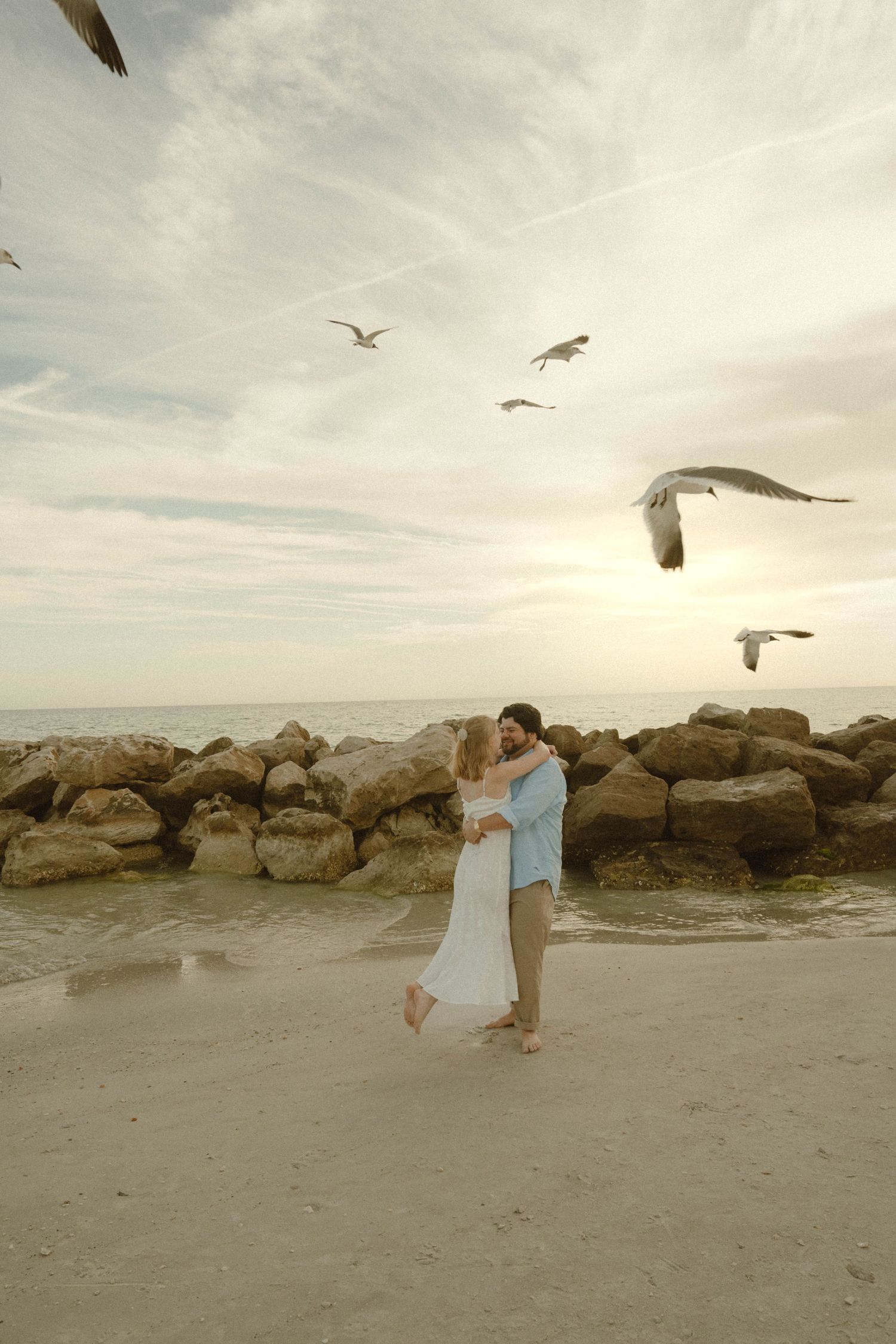 Couple embracing on beach surrounded by seagulls flying near rocky jetty at sunset.