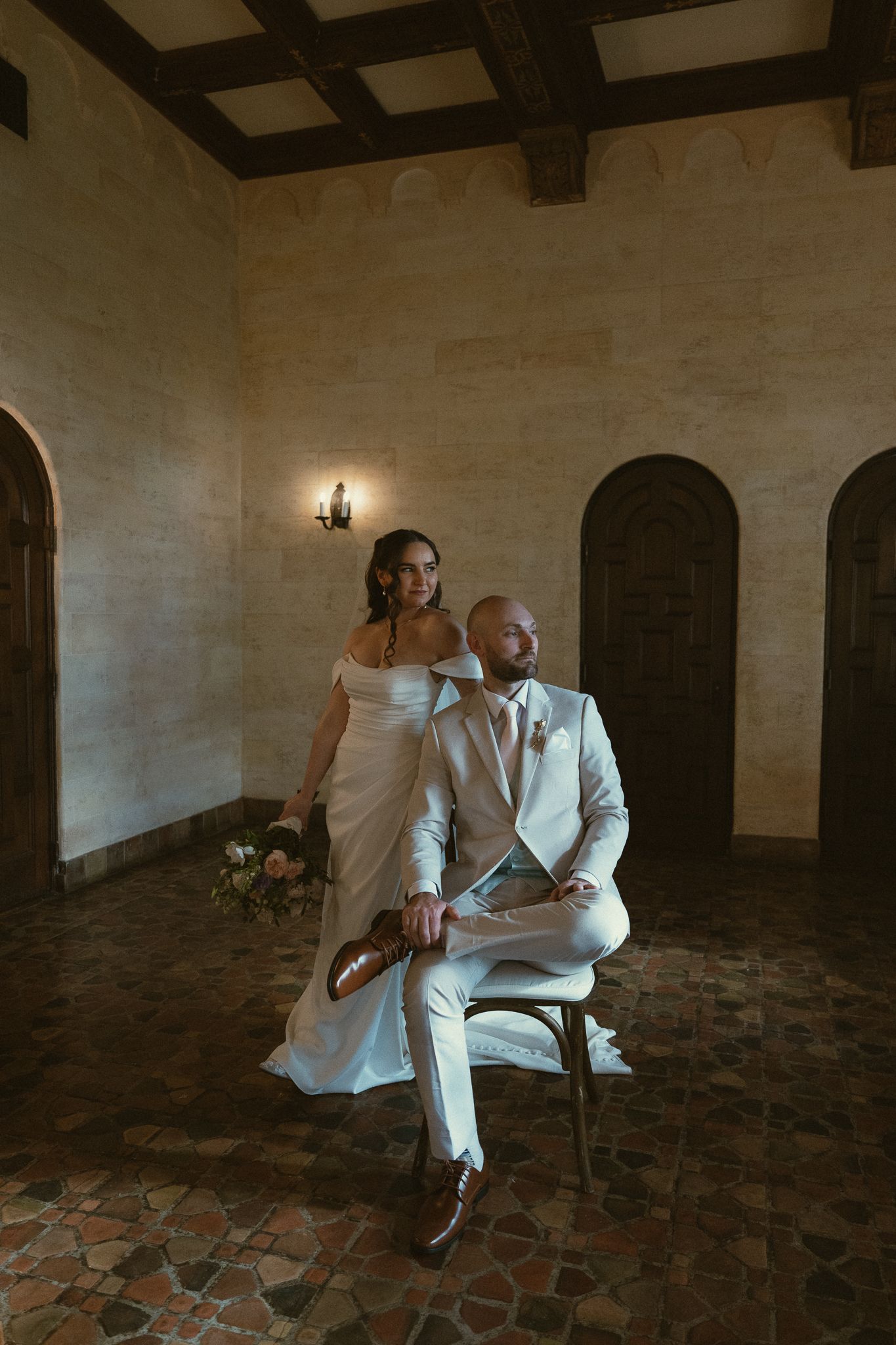 Bride in white off-shoulder gown stands behind seated groom in ivory suit inside elegant stone-walled venue.