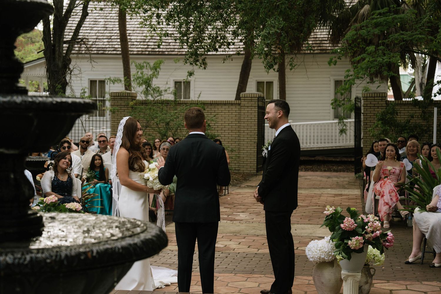 Outdoor wedding ceremony at a historic venue with a fountain, guests seated as couple exchanges vows.