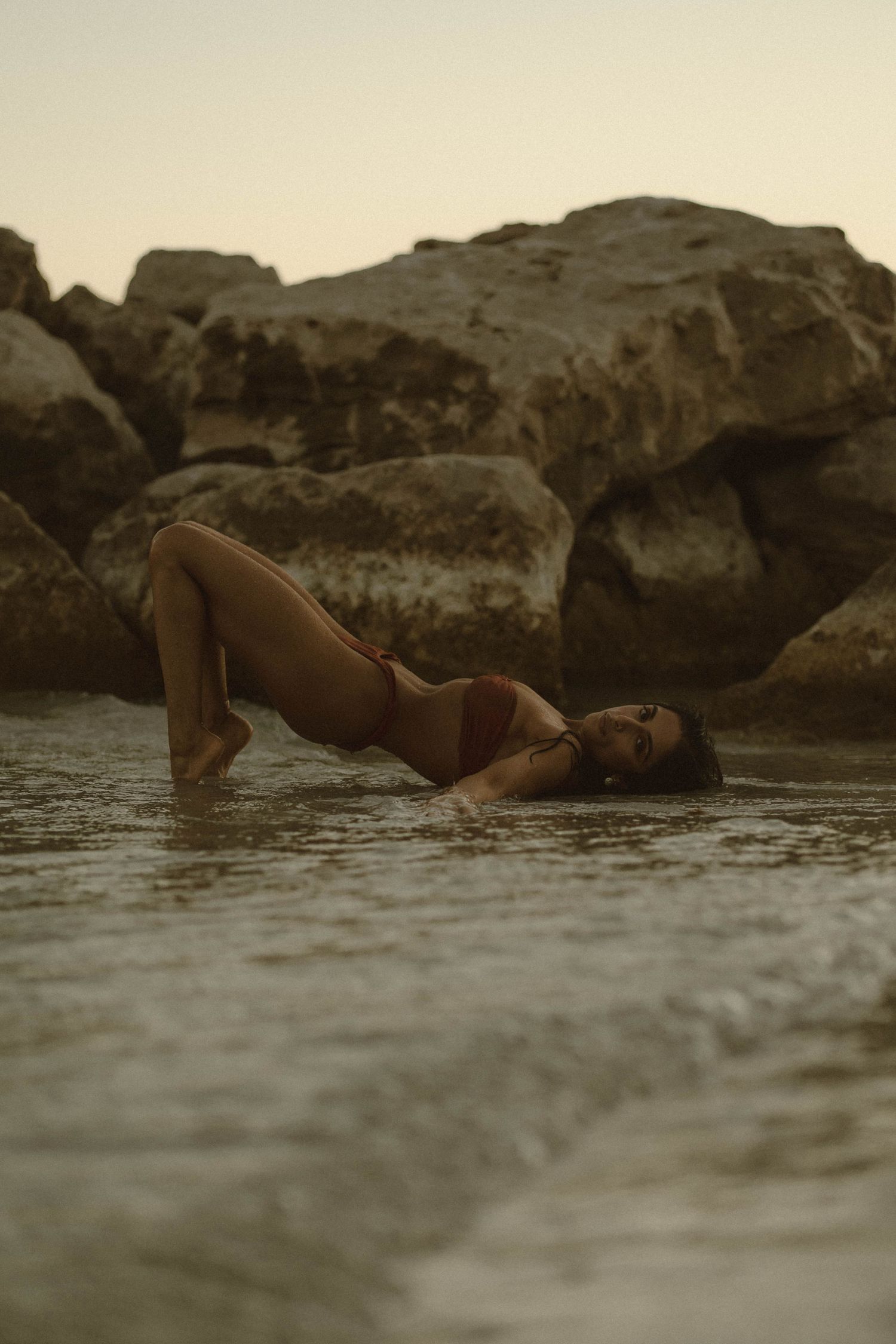 Woman in red bikini posing dramatically in shallow ocean water near large rocks at sunset.