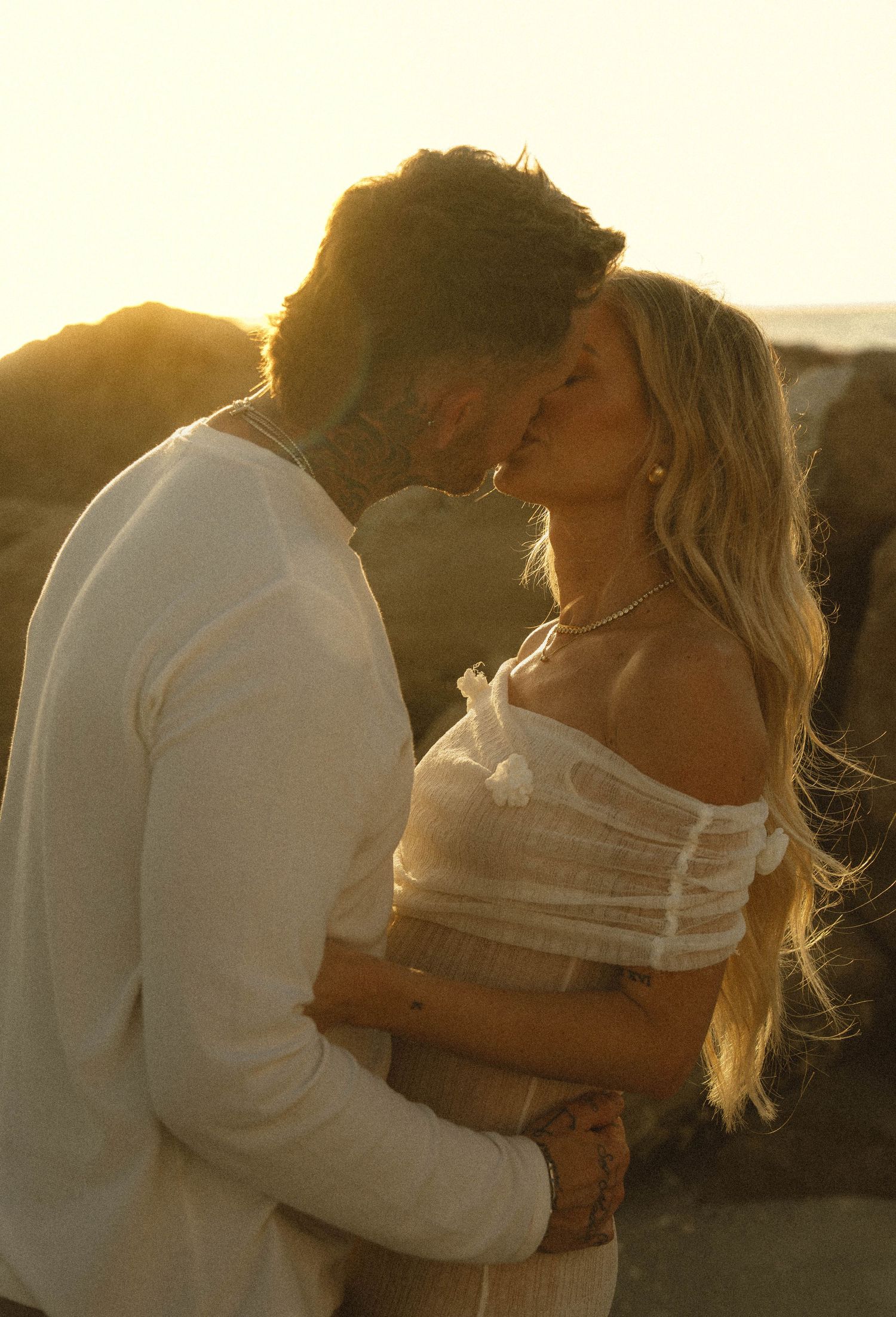A couple shares a golden-hour kiss on a rocky beach, bathed in warm sunset light.