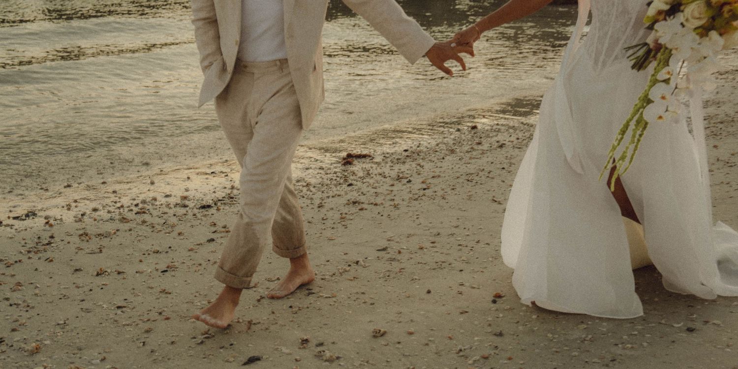 Barefoot newlyweds walk hand-in-hand along sandy beach, bride in white gown, groom in tan linen suit.