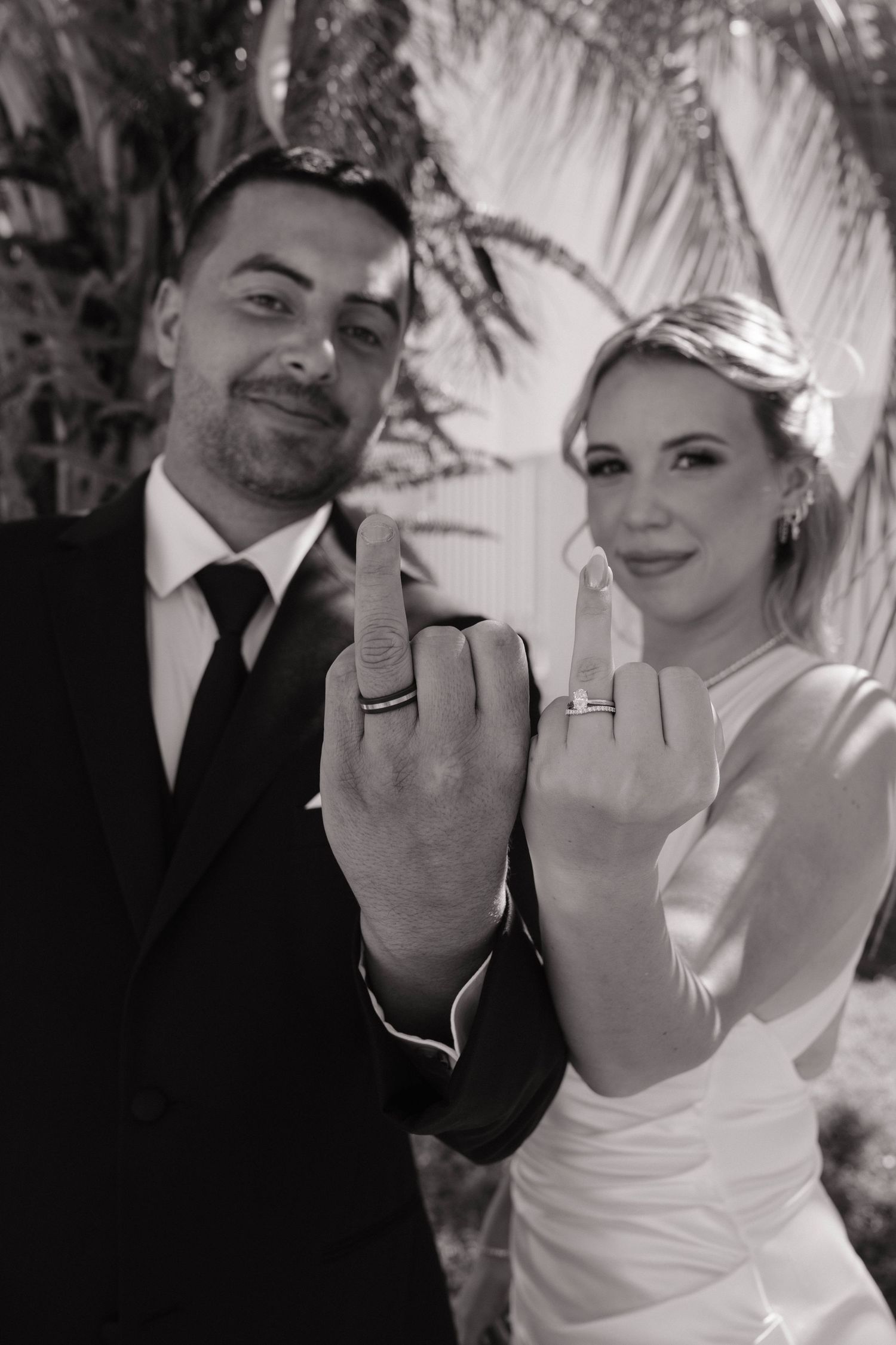 Black and white photo of smiling newlyweds showing off wedding rings on raised middle fingers.