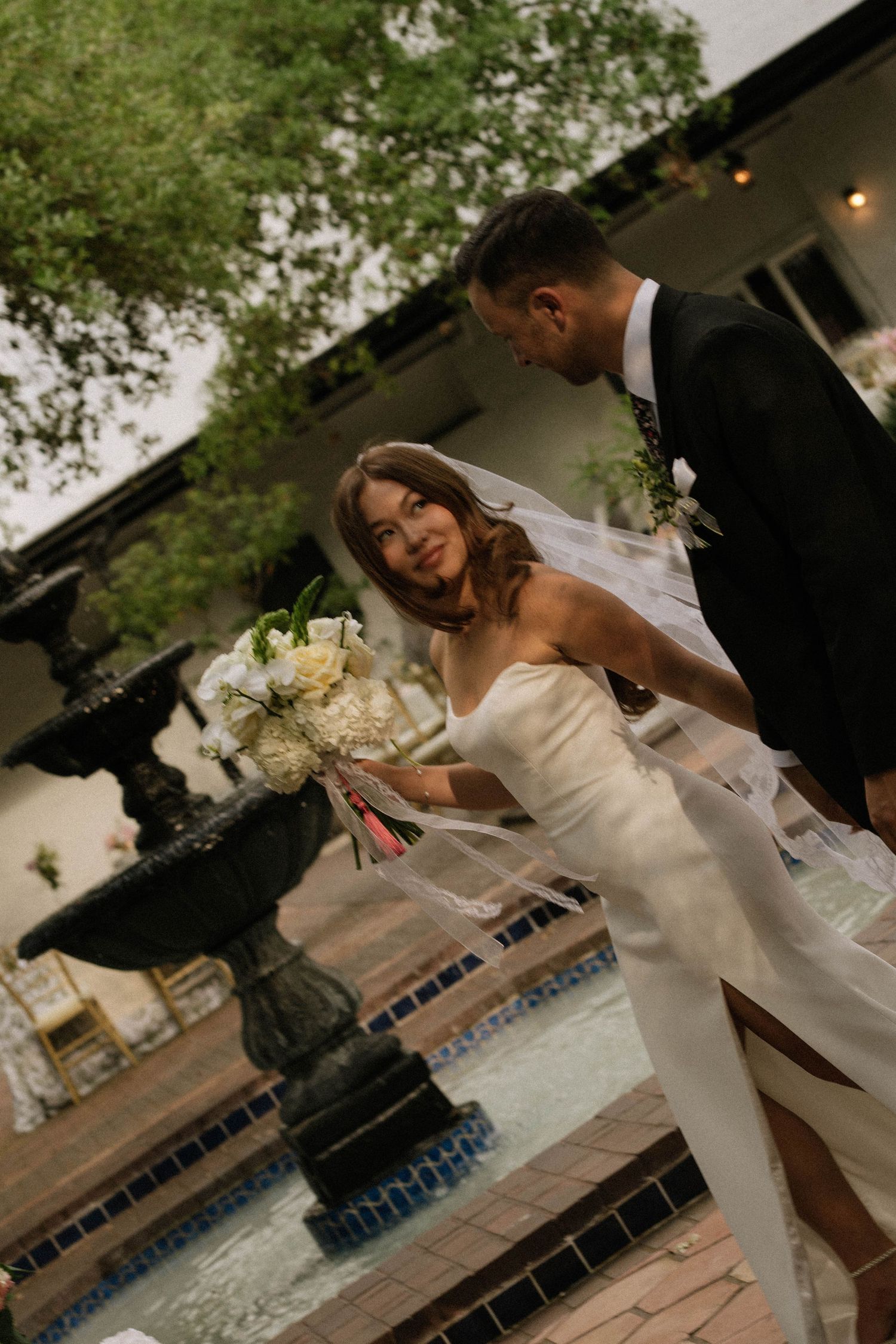 Bride in white gown with veil holding bouquet poses with groom in black suit near decorative fountain at dusk.