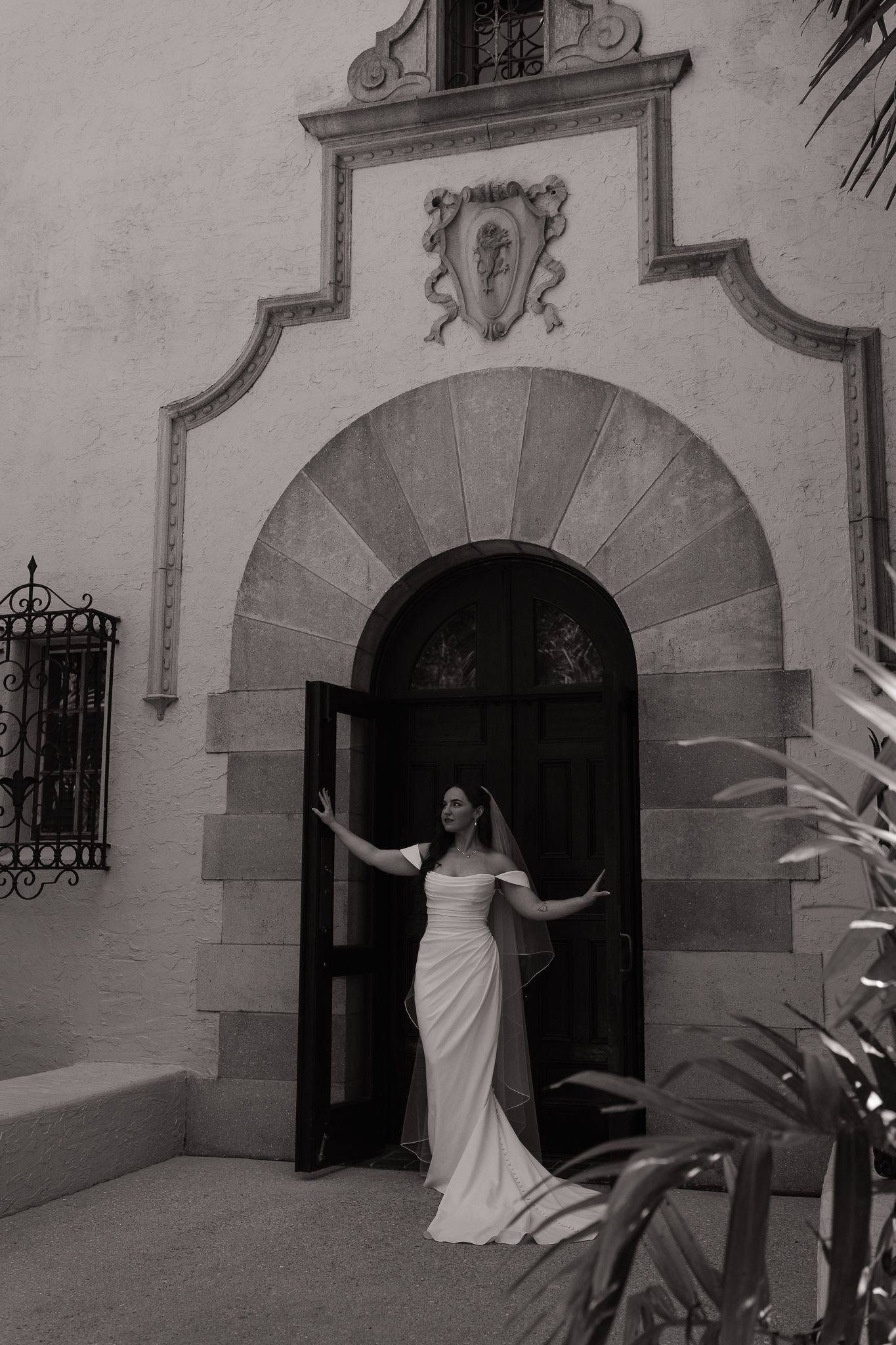 Bride in white gown poses at ornate arched doorway of historic building in black and white photo.