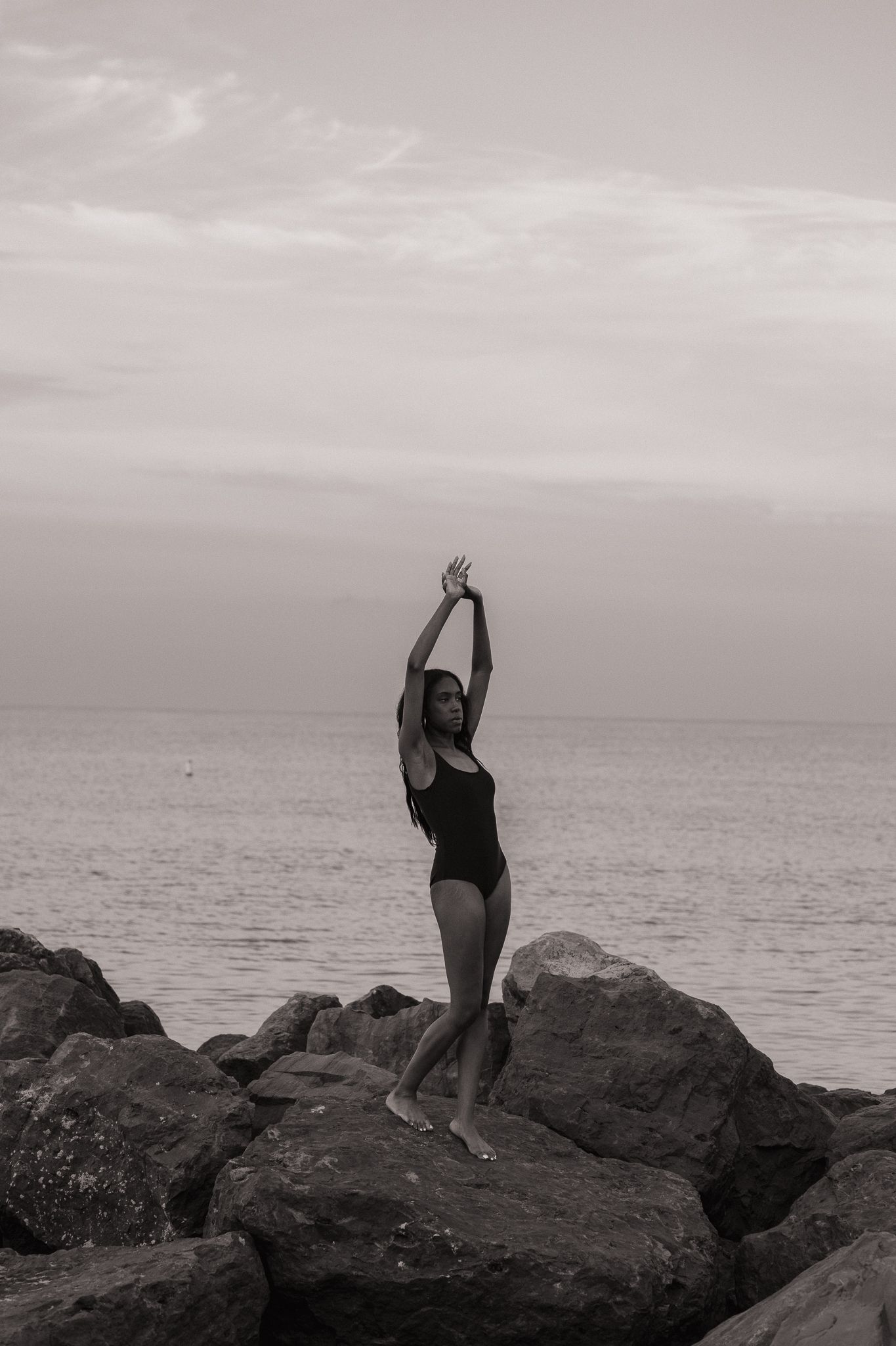 Woman in black swimsuit poses gracefully on rocks by the sea, arms raised, in a striking black and white photo.